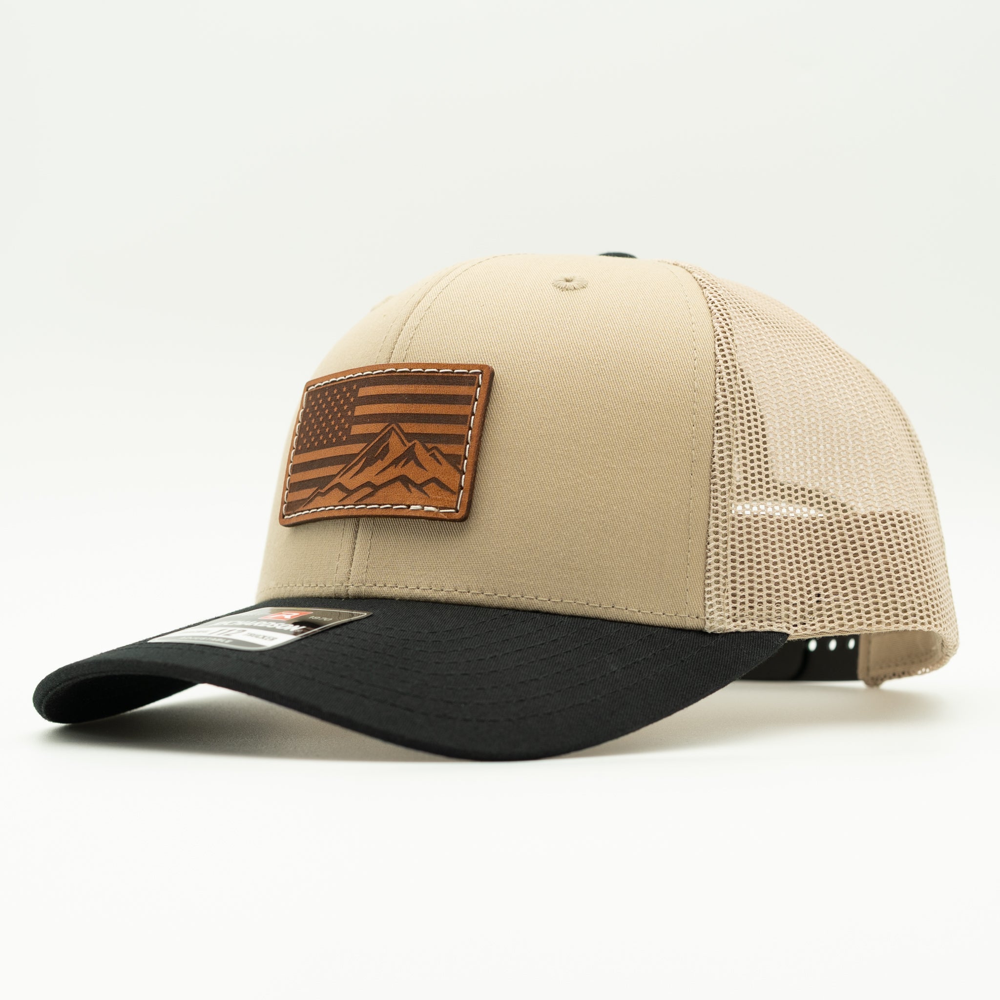 A structured trucker-style baseball cap with a beige front panel, black curved brim, and beige mesh back. It features a brown leather patch on the front with an American flag and mountain design.
Color: Khaki/Black Combo