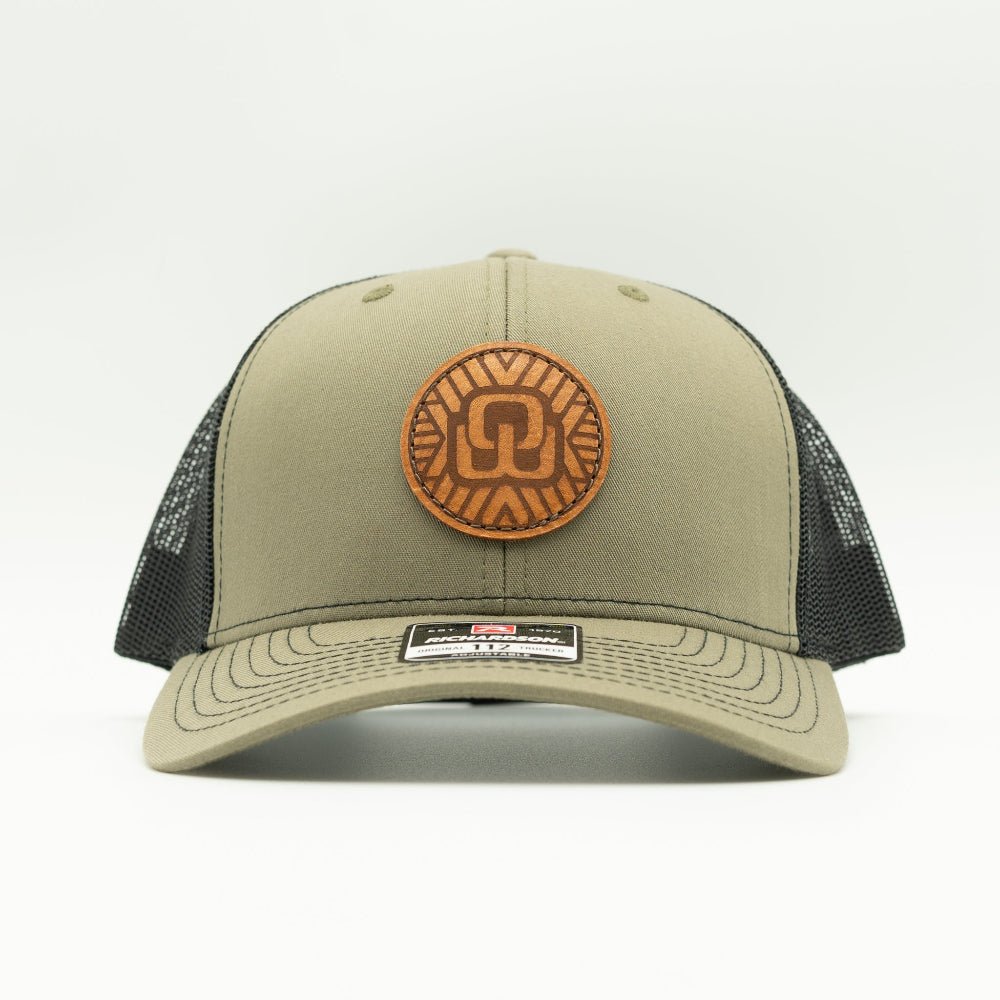 A trucker-style baseball cap in olive green with black mesh back panels and a circular brown leather patch on the front.
Color: Loden/Black