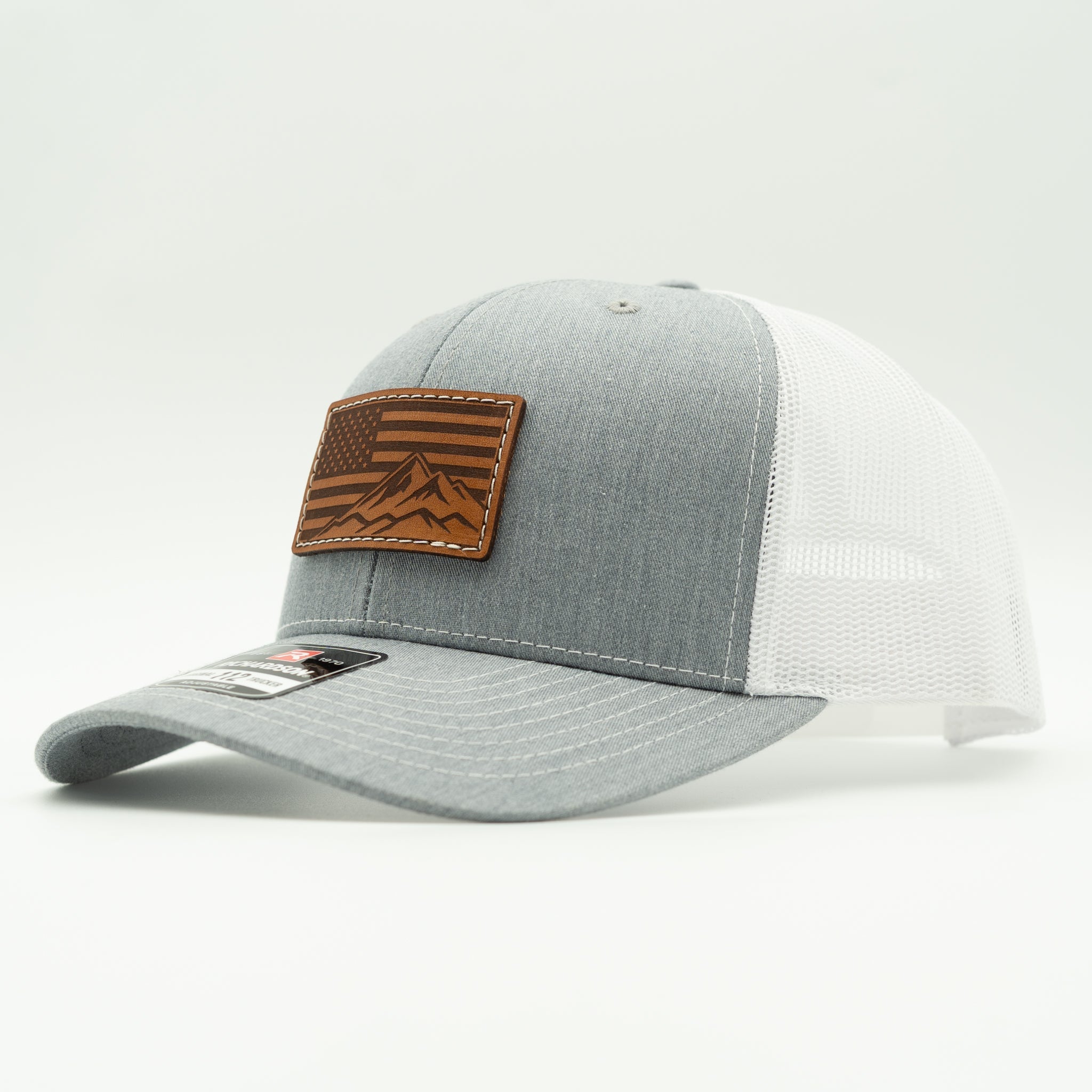 A two-tone trucker cap with a heather gray front panel and brim, white mesh back panels, and a brown leather patch featuring an American flag and mountain design on the front.
Color: Heather Grey/White