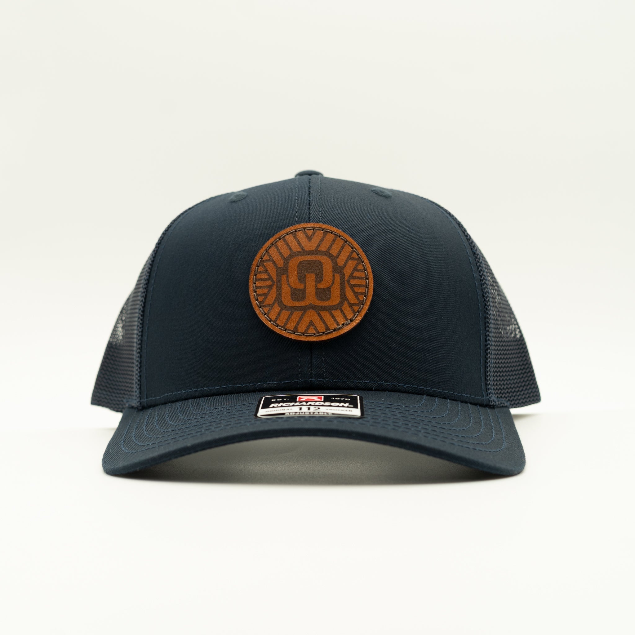 A dark navy blue trucker hat with a brown circular leather patch on the front featuring a stylized OW by geometric patterns. The hat has a structured front panel and a curved brim, with dark mesh back panels.
Color: Navy/Navy