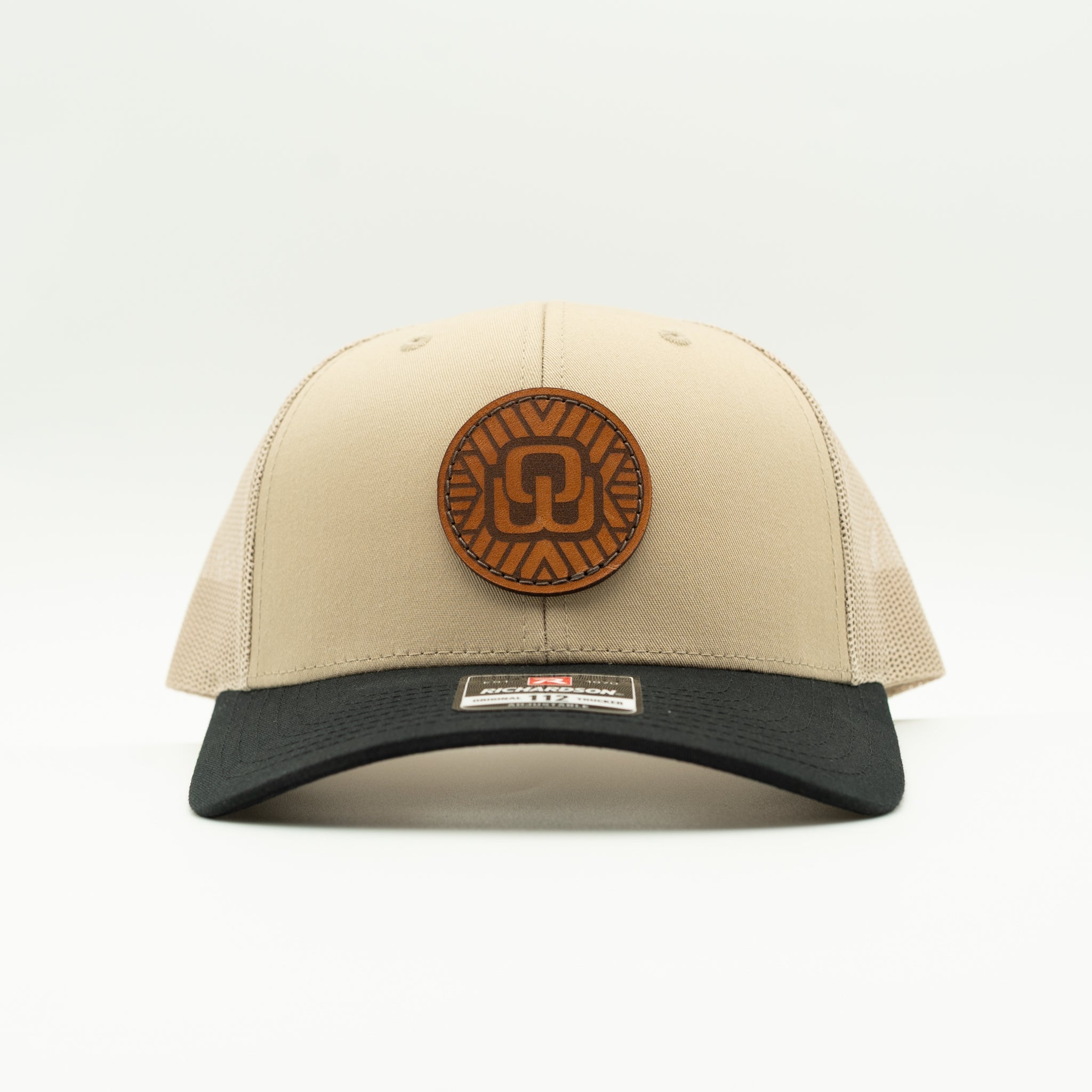 A structured trucker-style baseball cap with a beige front panel and mesh back, a black curved brim, and a circular brown leather patch on the front. A small rectangular brand label is visible on the brim.
Color: Khaki/Black Combo