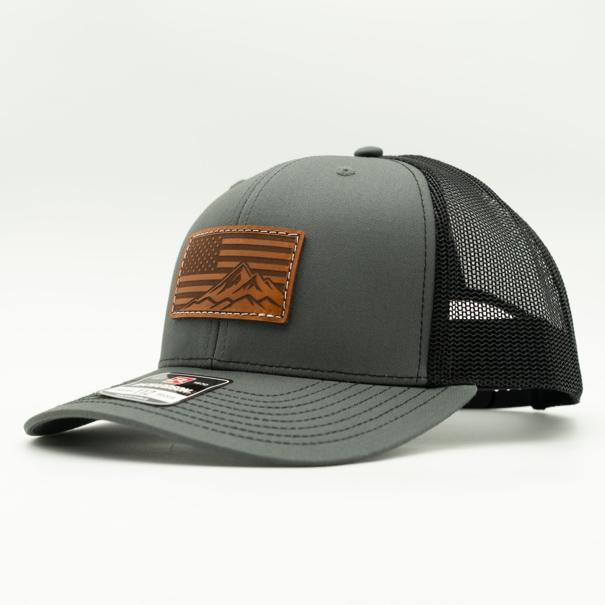 Side profile of American Range American flag leather patch hat on charcoal and black Richardson 112 trucker, showing patch, curved bill, mesh back, and snapback, a casual patriotic mountain hat for outdoor lifestyles and daily use.
Color: Charcoal/Black