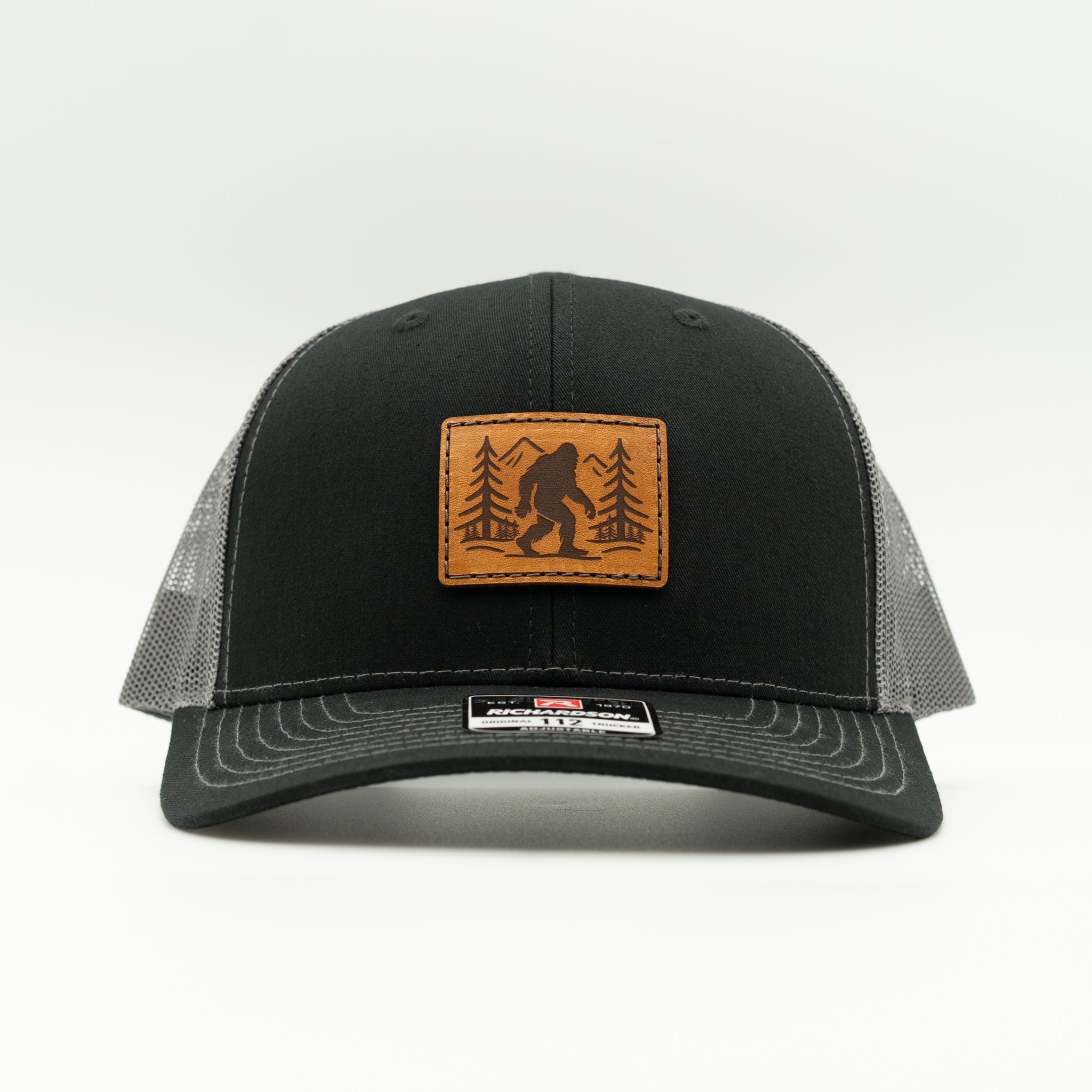 Front view of black/charcoal Mountain Myth Bigfoot leather patch trucker hat, structured mid-profile Richardson 112 with mesh back.