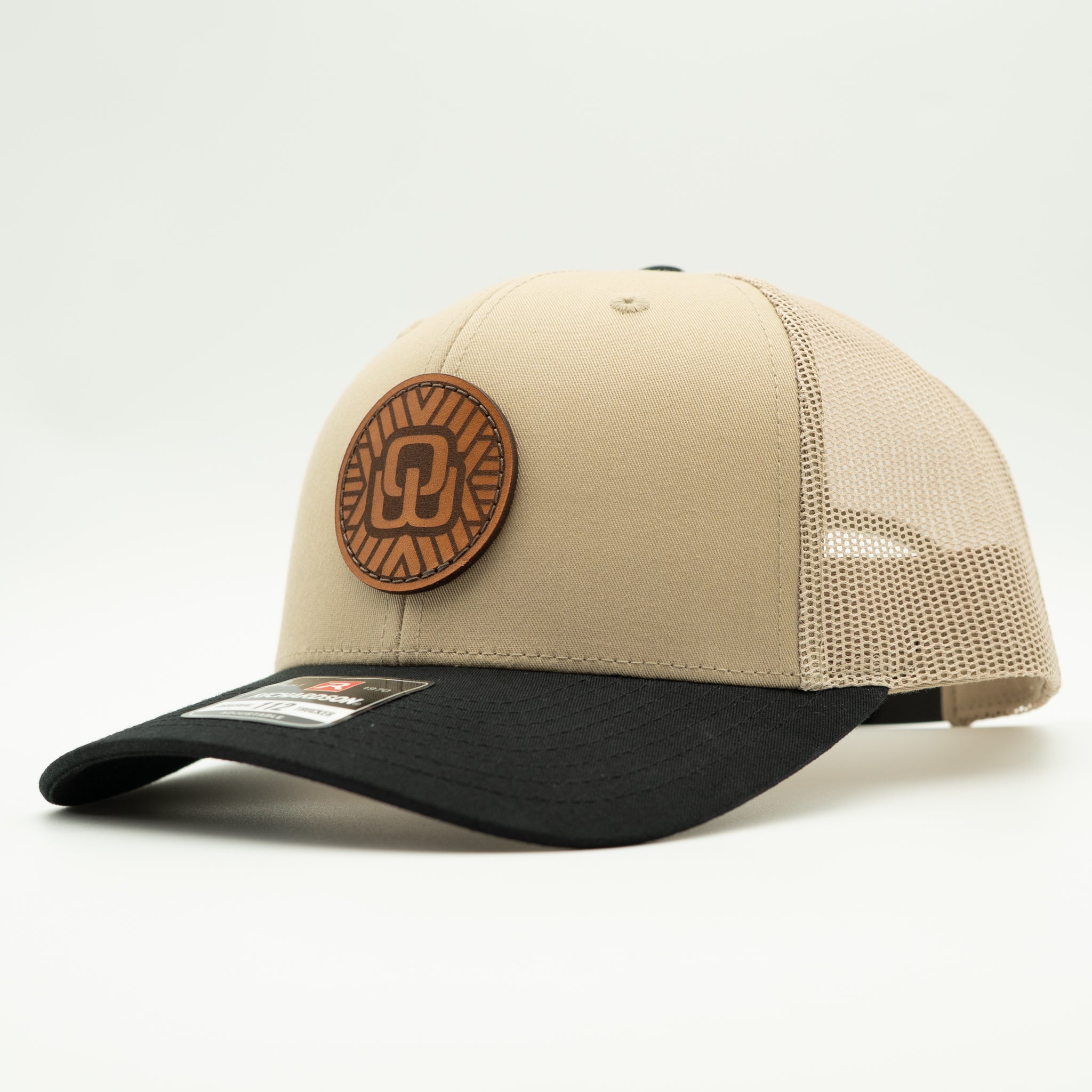 A beige and black trucker-style baseball cap with a circular brown leather patch on the front, photographed on a clean white background.
Color: Khaki/Black Combo