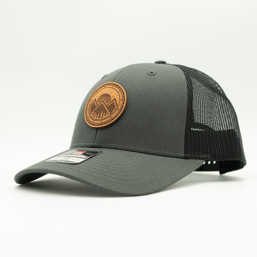 A dark gray trucker-style cap with black mesh back panels and a circular brown leather patch on the front. The patch features a stylized mountain logo.
Color: Charcoal/Black