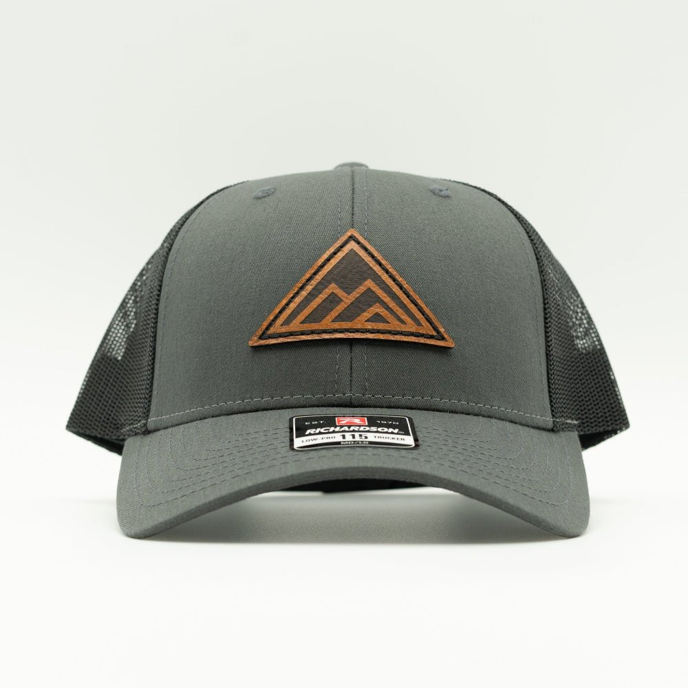 Dark Mountain leather patch hat on a charcoal and black Richardson 115 low profile trucker hat, front view showing triangle mountain patch, curved bill, mesh back, and snapback, a low profile mountain hat for everyday outdoor wear.
Color: Charcoal/Black