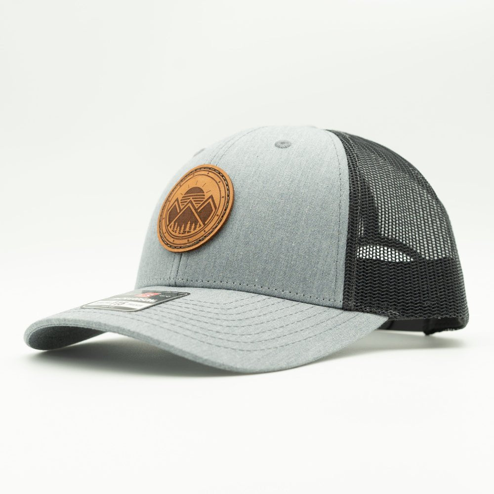 A light gray and black trucker hat with a brown circular leather patch on the front, photographed on a clean white background.
Color: Heather Grey/Charcoal