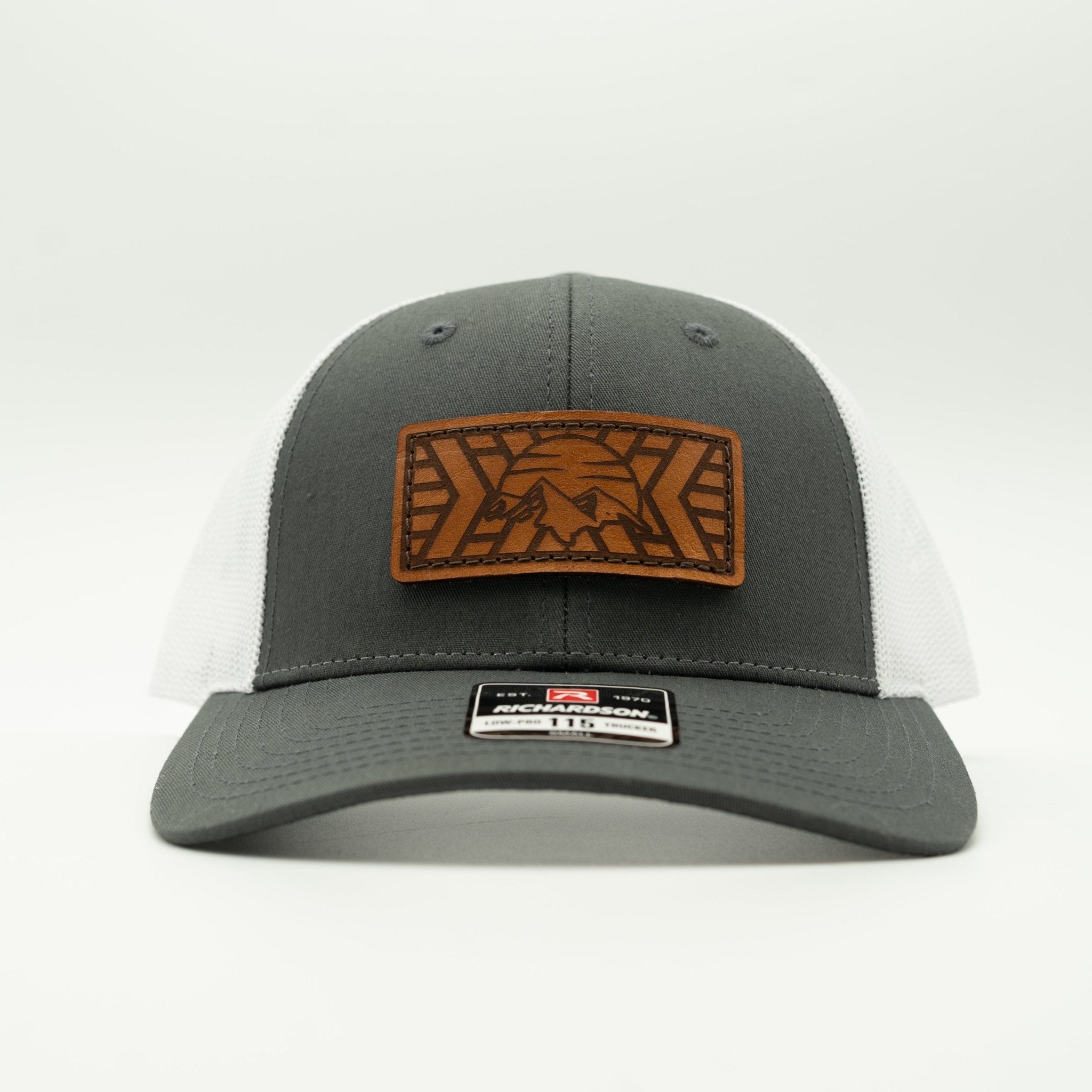 A dark gray and white trucker hat with a brown leather patch on the front featuring a mountain design. The hat has a dark gray front panel and visor, white mesh back.
Color: Charcoal/White
