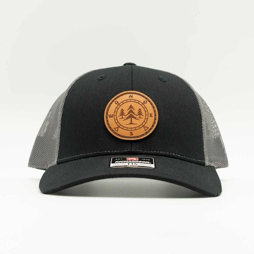 A black trucker hat with a circular brown leather patch on the front featuring a tree design and compass directions. The back panels are made of gray mesh.
Color: Black/Charcoal