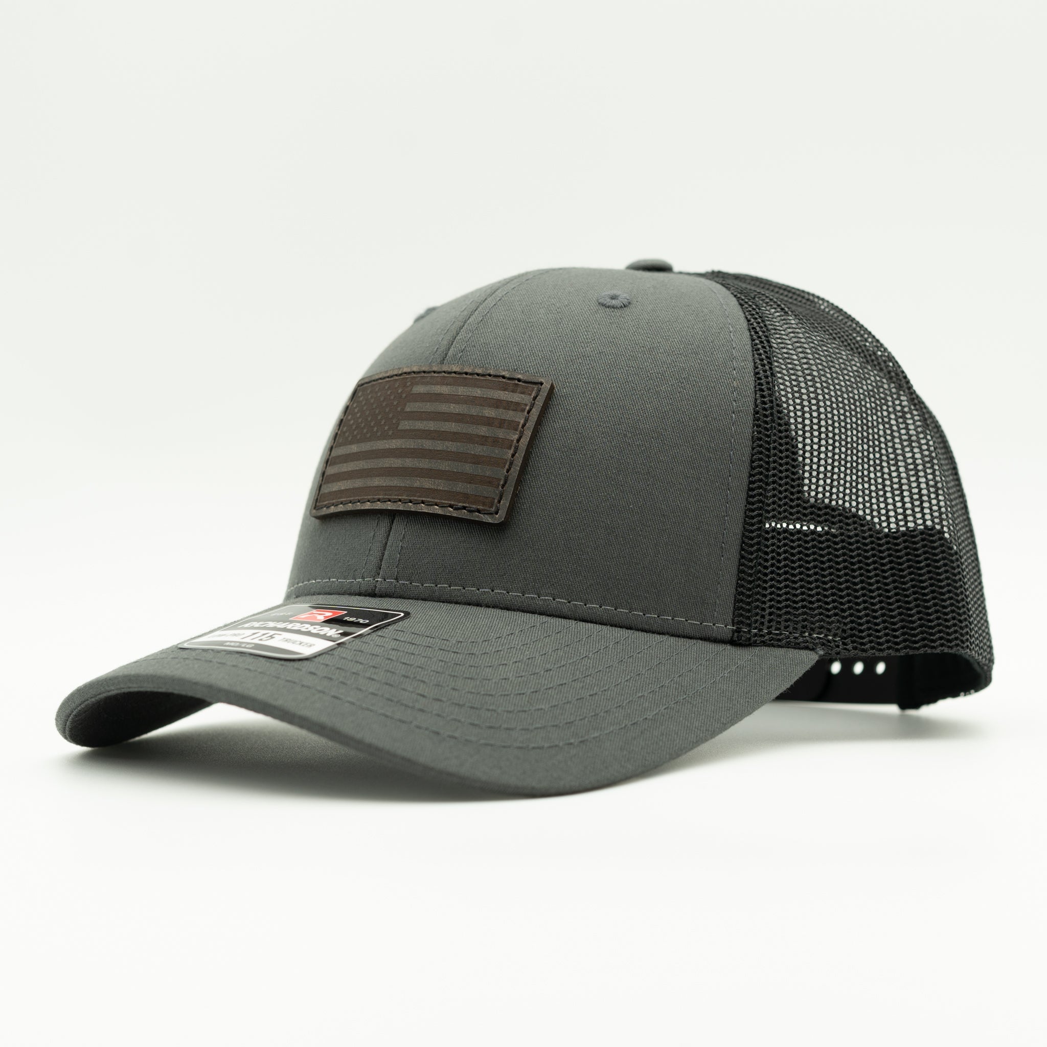A dark gray and black trucker cap with a brown leather American flag patch on the front, shown on a light gray background.
Color: Charcoal/Black