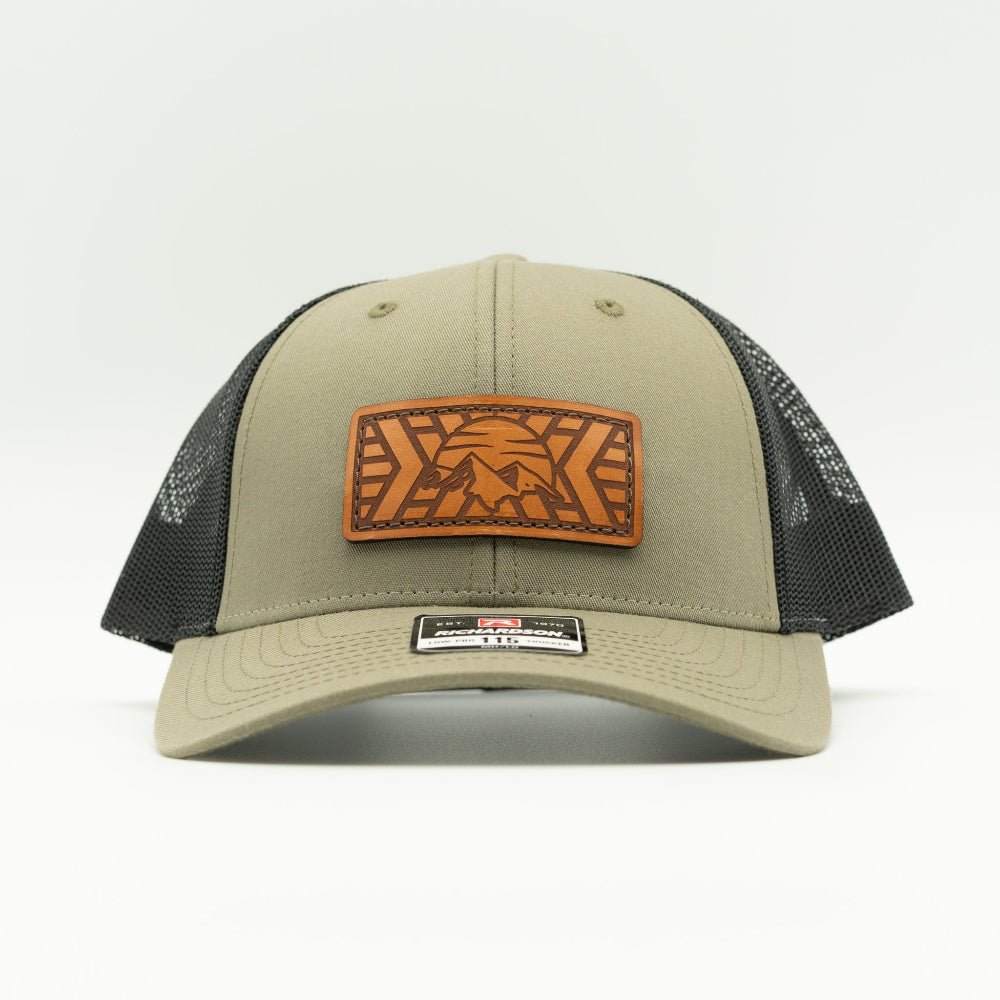 A trucker-style baseball cap in loden green with black mesh back panels and a brown leather patch on the front featuring a stylized eagle design.
Color: Loden/Black
