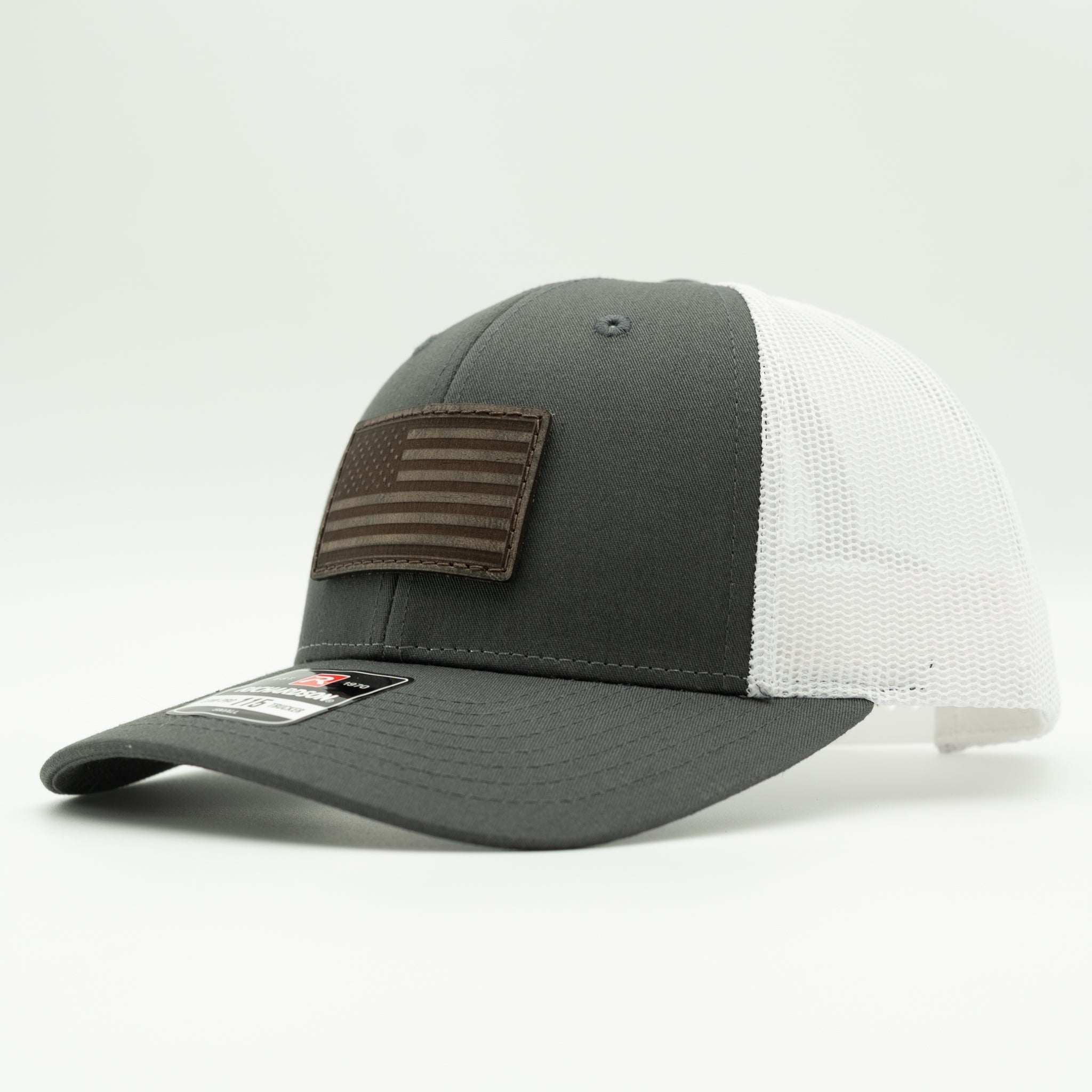 A dark gray and white trucker-style baseball cap with a brown leather American flag patch on the front, photographed on a clean white background.
Color: Charcoal/White