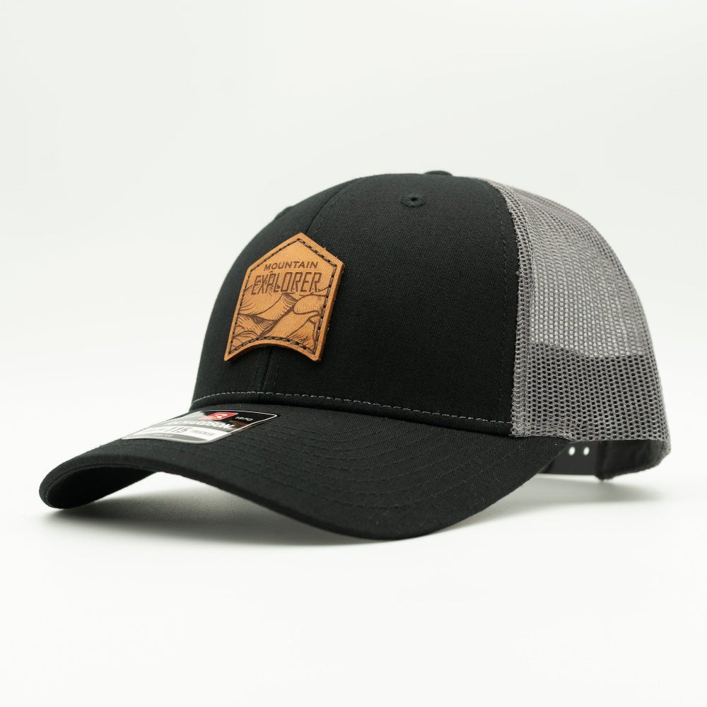 Side angle of Mountain Explorer leather patch hat on black and charcoal Richardson 115 low profile trucker, highlighting two-tone crown, mesh back, and curved bill.
