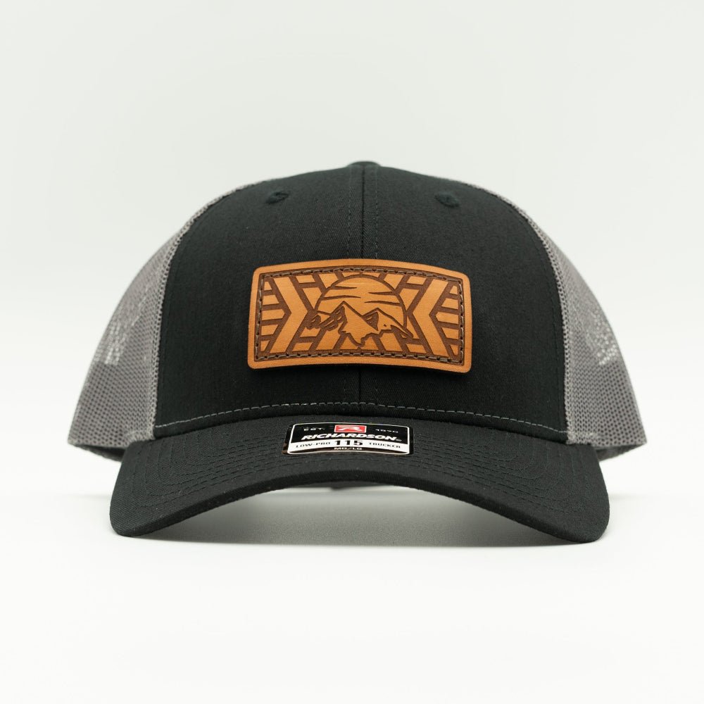 A black trucker hat with gray mesh back panels and a brown leather patch on the front featuring a mountain and sun
design.
Color: Black/Charcoal
