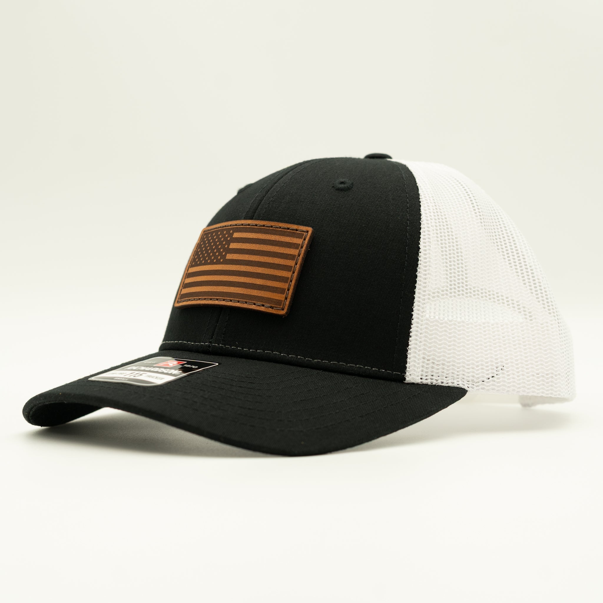 A black and white trucker hat with a brown leather American flag patch on the front, photographed on a clean white background.
Color: Black/White