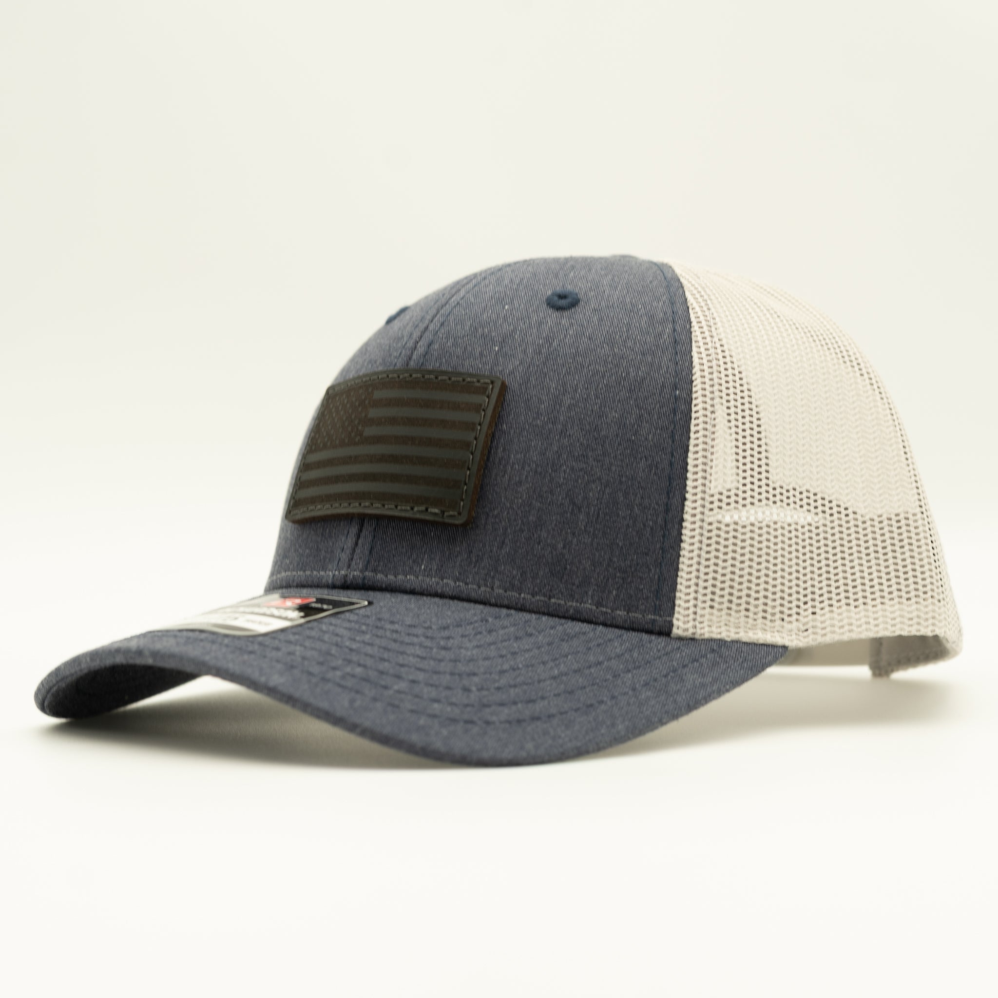 A trucker-style baseball cap with a dark heather gray front panel and brim, off-white mesh back panels, and a dark olive green rectangular patch on the front depicting a stylized American flag.
Color: Heather Navy/Grey