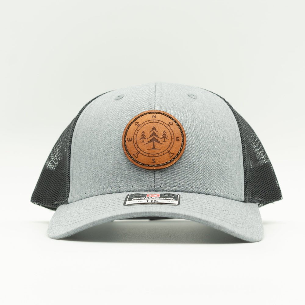 A light gray and charcoal mesh trucker hat with a circular brown leather patch on the front featuring three pine trees, photographed against a clean white background.
Color: Heather Grey/Charcoal
