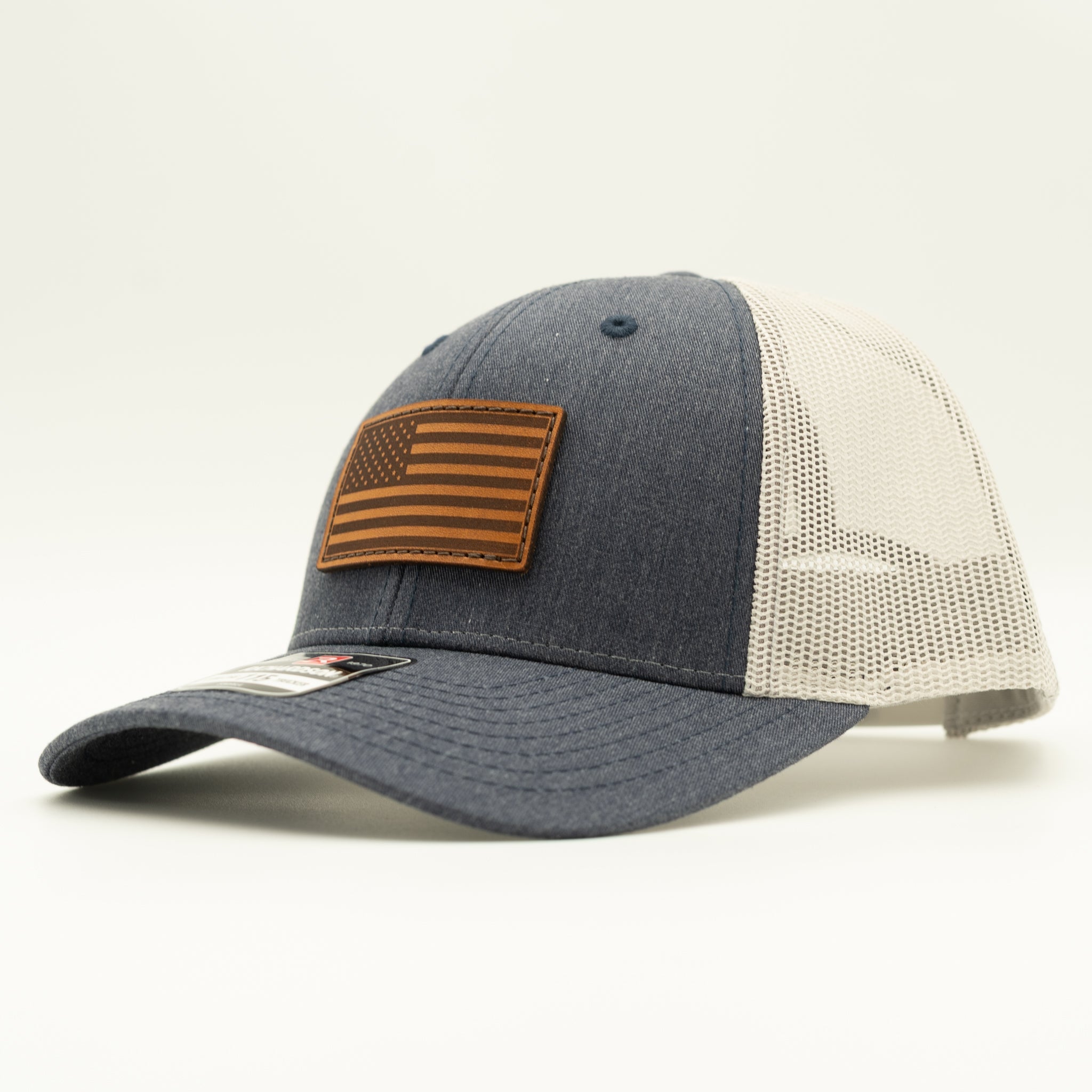 A trucker-style baseball cap with a dark gray fabric front and brim, off-white mesh back panels, and a brown leather American flag patch on the front.
Color: Heather Navy/Grey