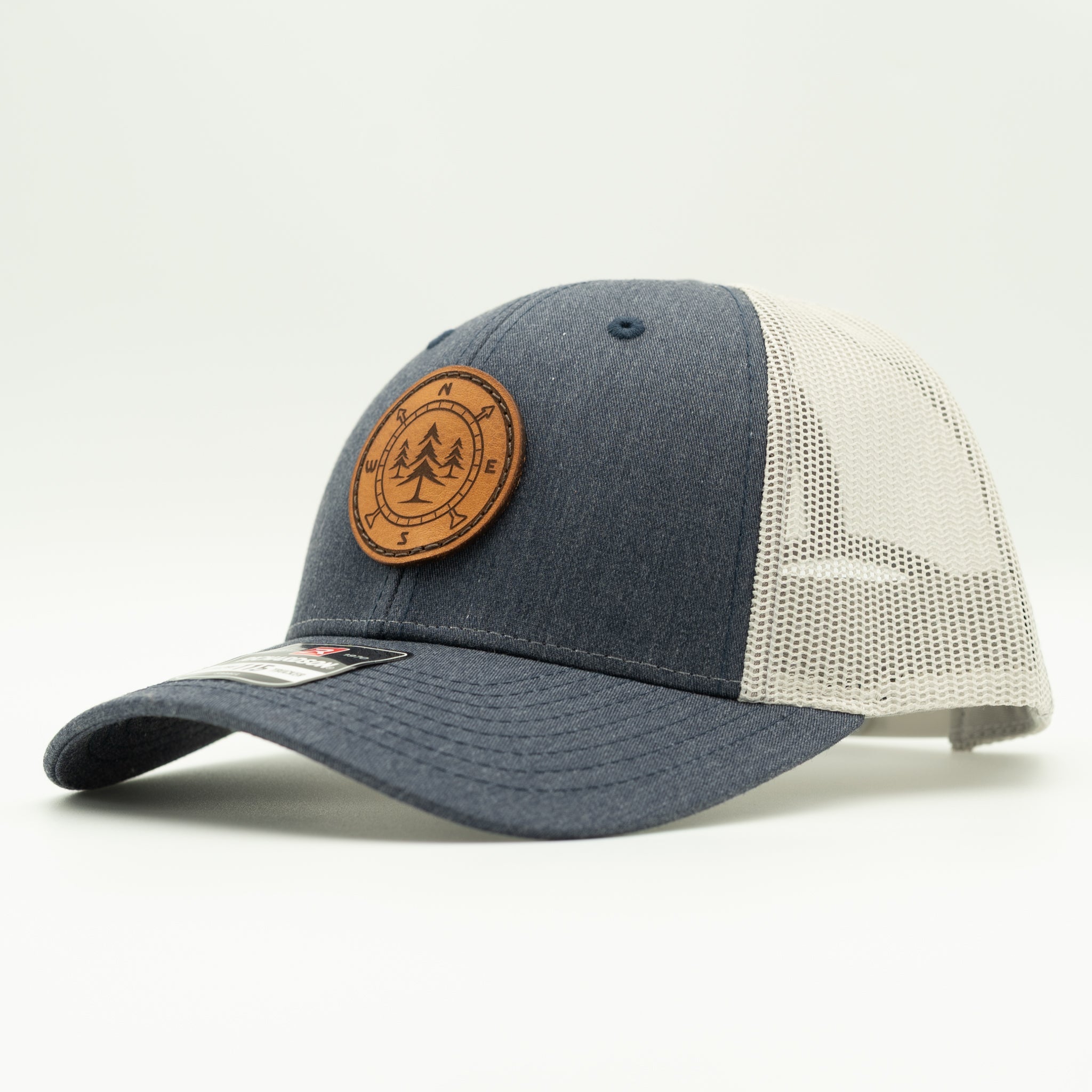 A trucker-style baseball cap with a heather navy blue front panel and brim, grey mesh back panels, and a circular brown leather patch on the front featuring a pine tree and compass laser engraving.
Color: Heather Navy/Grey