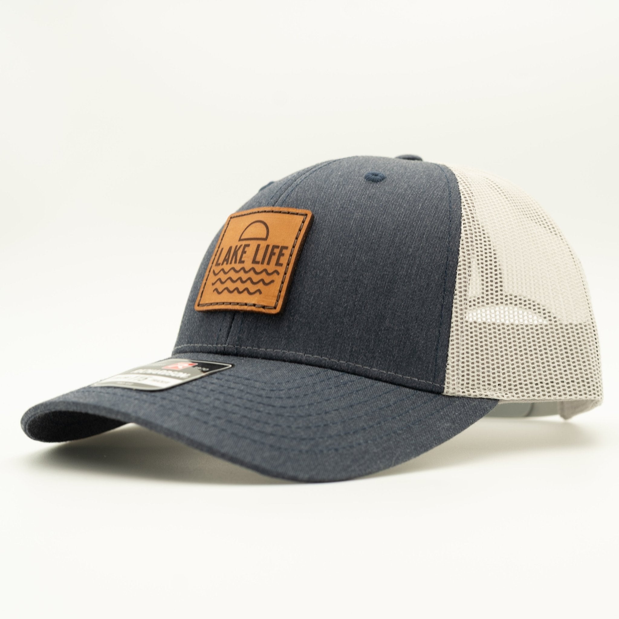 A trucker-style baseball cap with a dark navy blue front panel and brim, off-white mesh back panels, and a brown leather patch on the front that reads 'LAKE LIFE'.
Color: Heather Navy/Grey