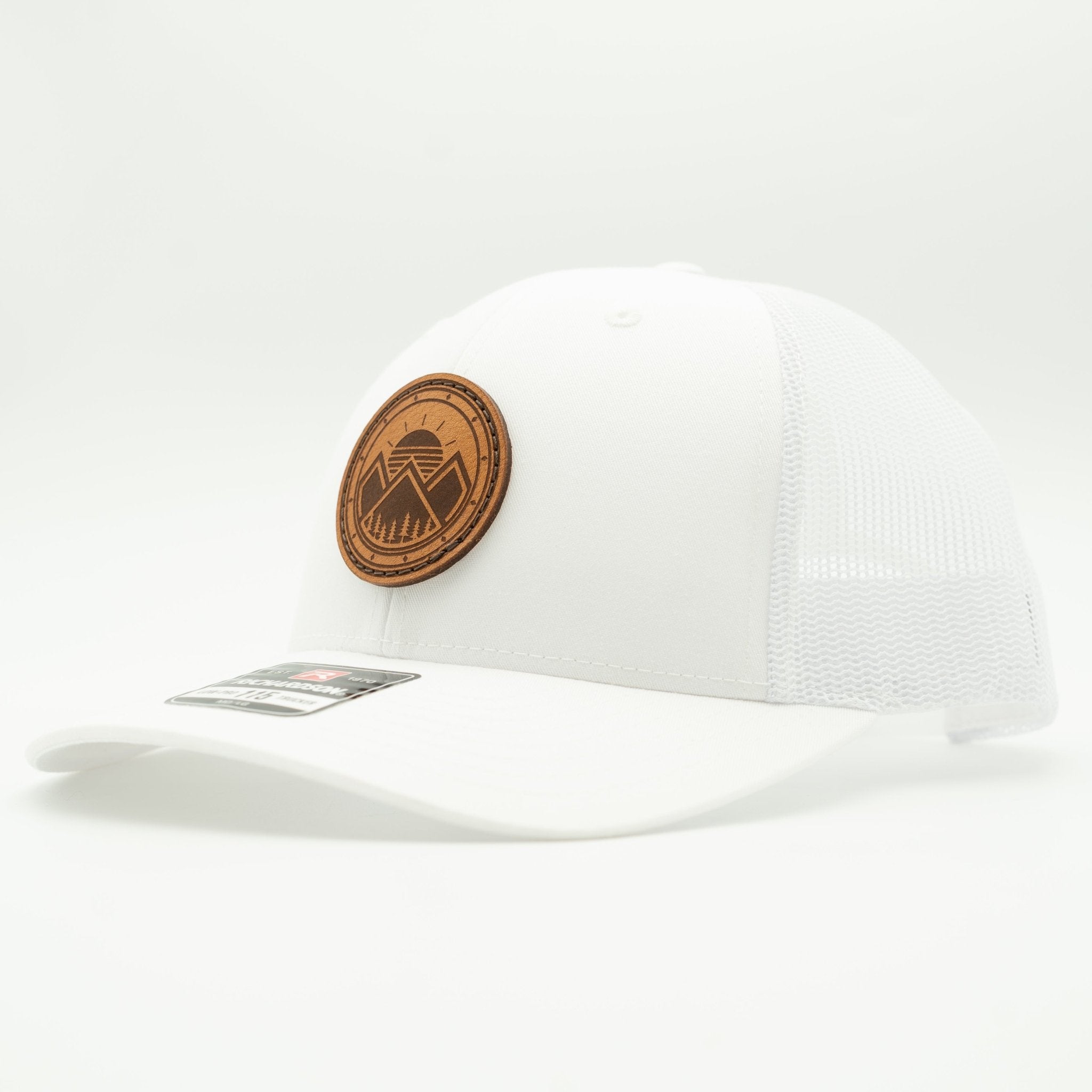 A white trucker-style baseball cap with a curved brim, mesh back panels, and a circular brown leather patch on the front featuring a mountain and tree design. A small retail sticker is visible on the brim.
Color: Solid White