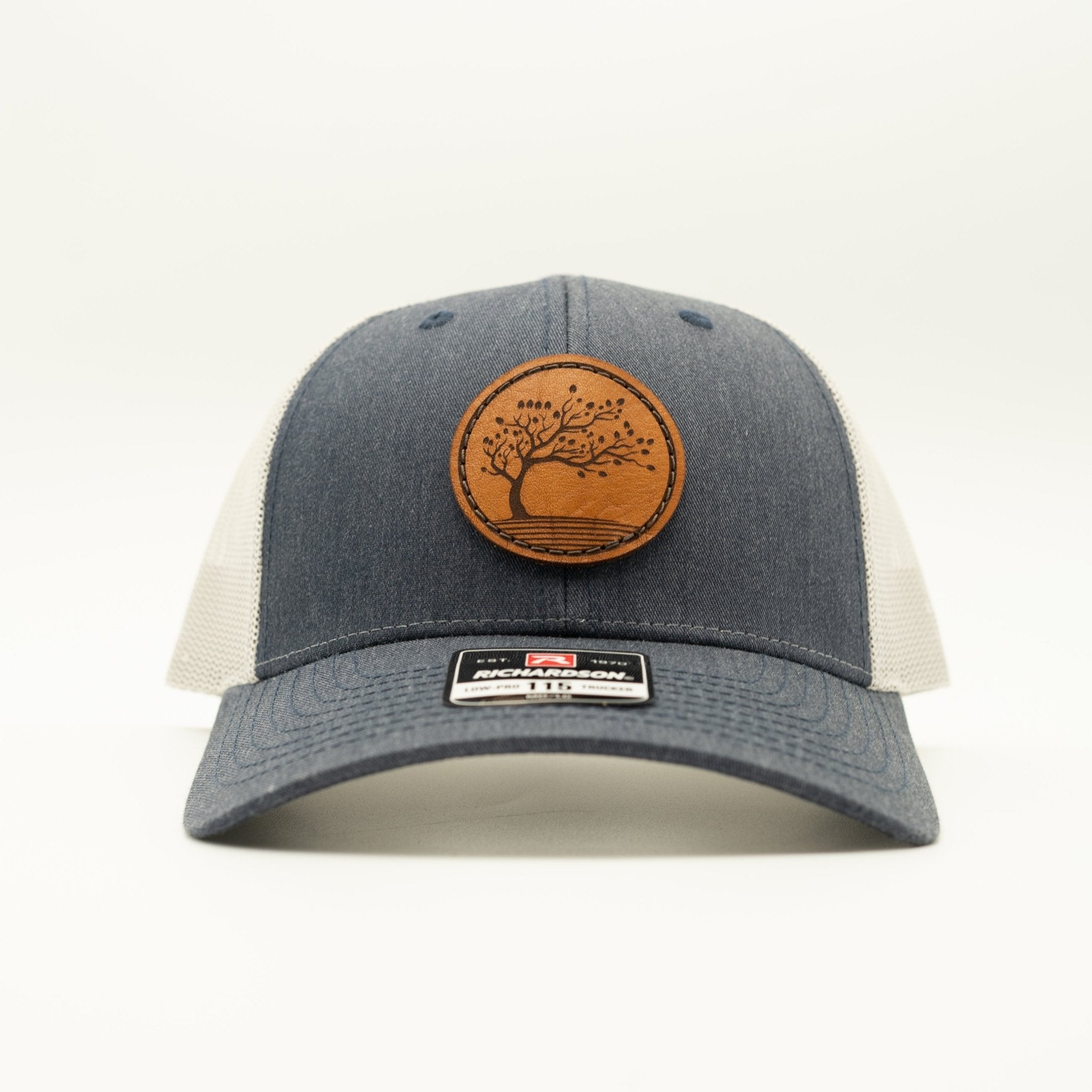 A trucker-style baseball cap with a dark gray heather front and brim, off-white mesh back panels, and a circular brown leather patch featuring a tree design on the front. A small rectangular brand label is visible on the brim.
Color: Heather Navy/Grey
