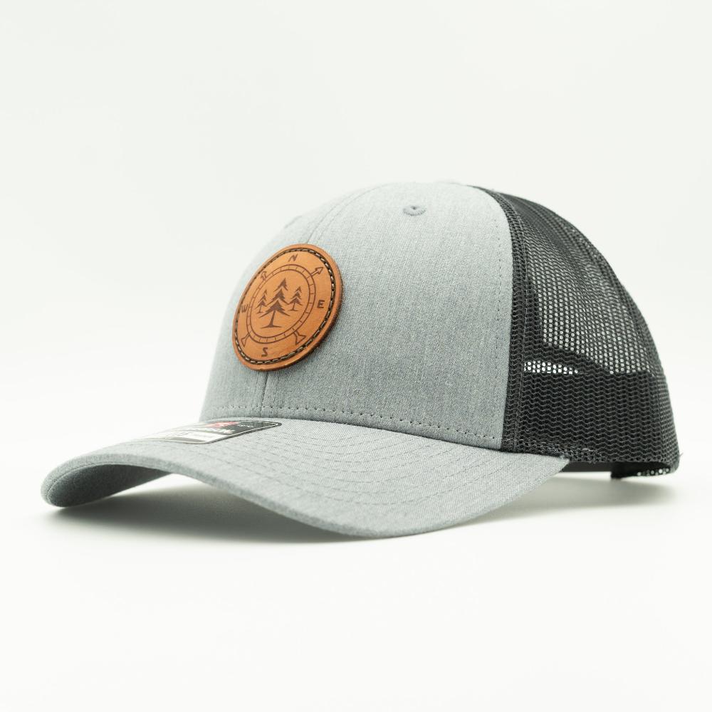 A light gray and black trucker-style baseball cap with a curved brim, dark charcoal mesh back, and a circular brown leather patch on the front featuring a pine tree and compass engraving.
Color: Heather Grey/Charcoal