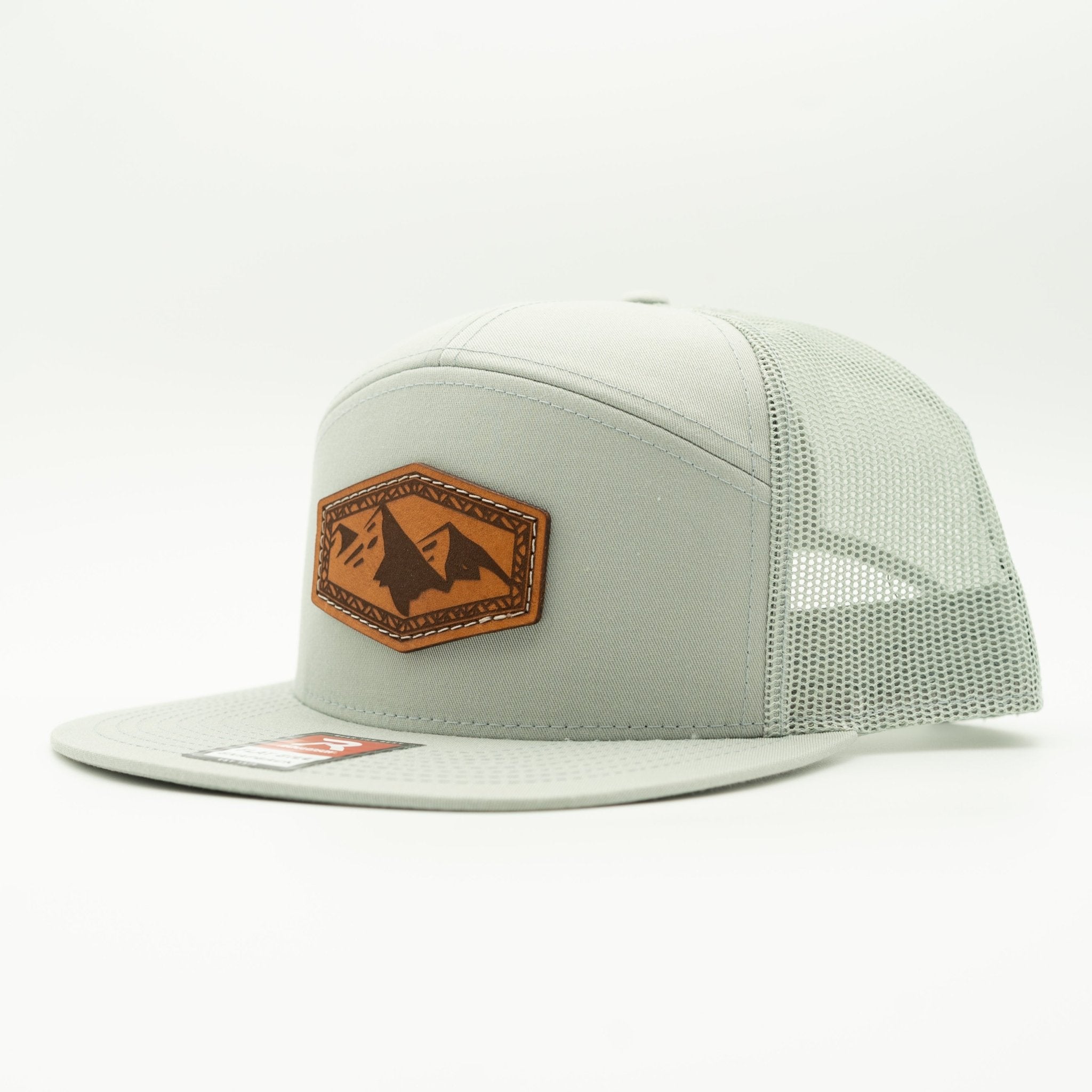 A light gray trucker cap with a structured front panel, matching flat brim, and mesh back panels. It features a brown leather patch on the front with a mountain logo, and a small red and white brand sticker on the brim.
Color: Quarry 168 (OSFM)