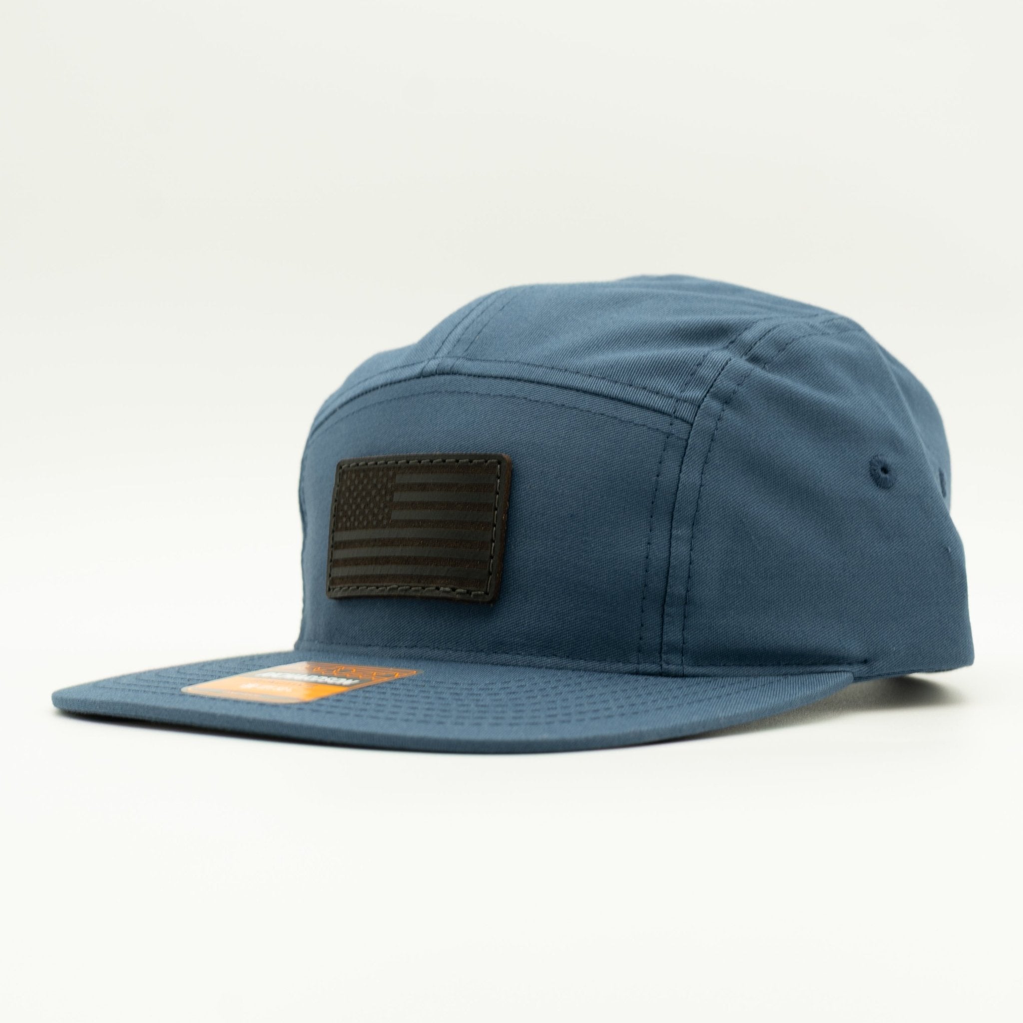 A blue five-panel cap with a dark brown leather American flag patch on the front and a matching blue flat brim, photographed on a clean white background.
Color: Light Navy