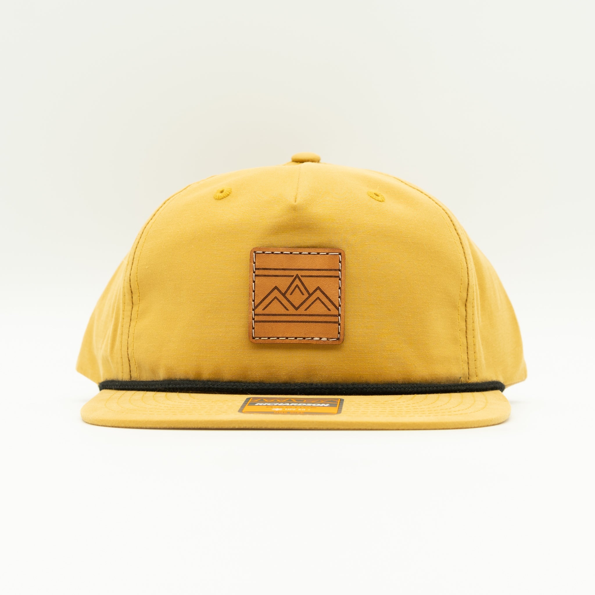 Rustic Ridge biscuit and black Richardson 256 rope hat front view with light brown leather mountain patch on tan crown and black flat bill.