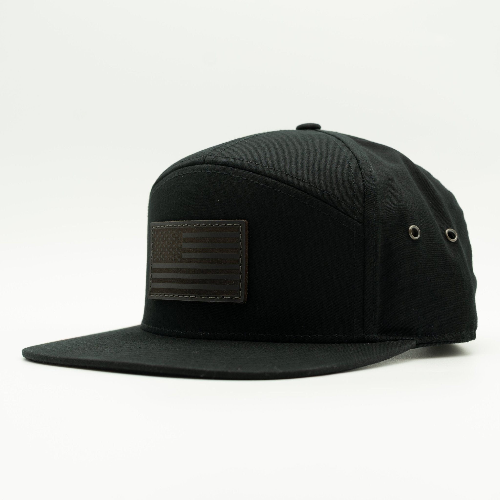 A black flat-brim cap with a dark brown rectangular American flag patch on the front, photographed on a clean white background.
Color: Black