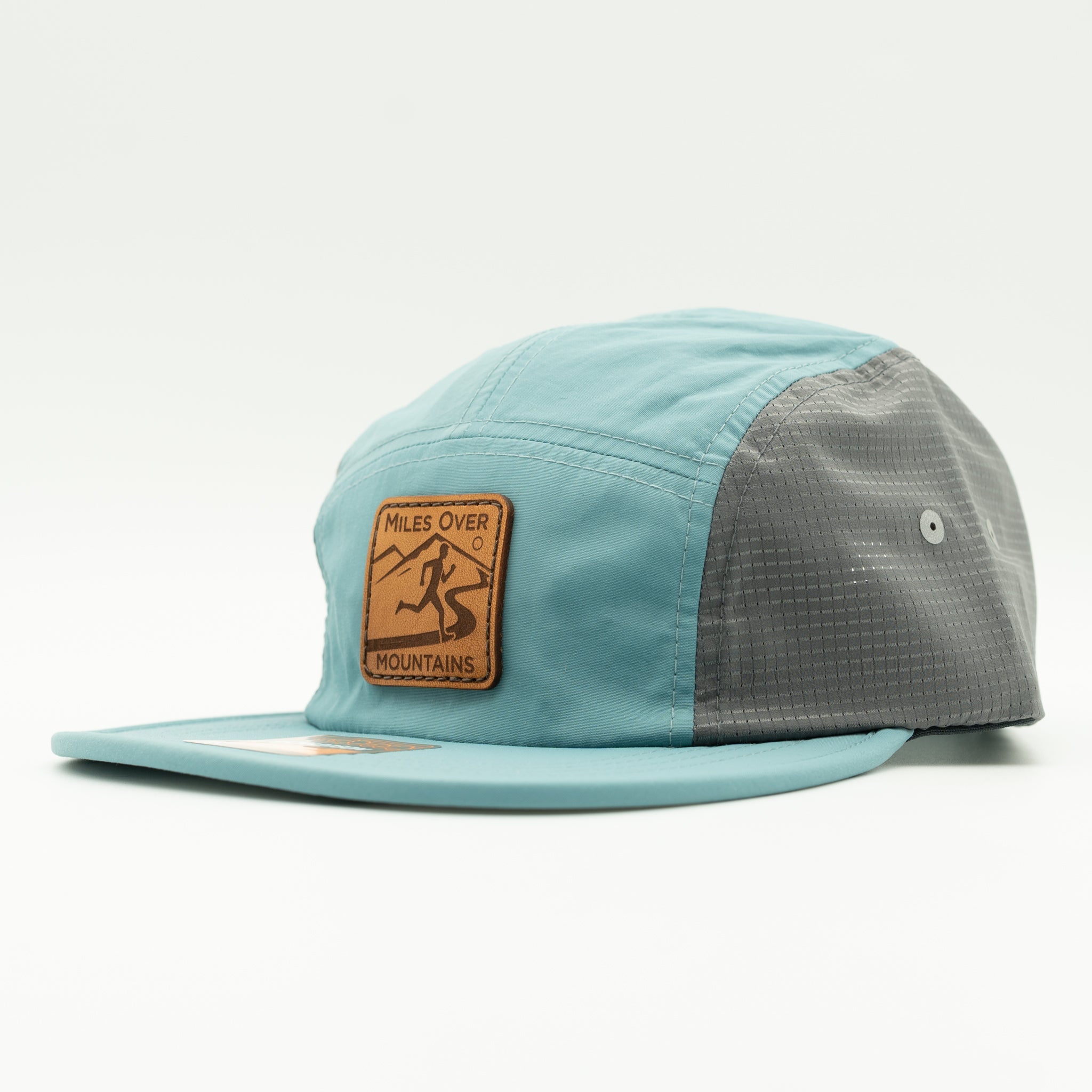 A 5-panel camp cap by Miles Over Mountains in light blue and dark gray, featuring a brown leather patch on the front with a mountain and skier logo.
Color: Smoke Blue/Charcoal