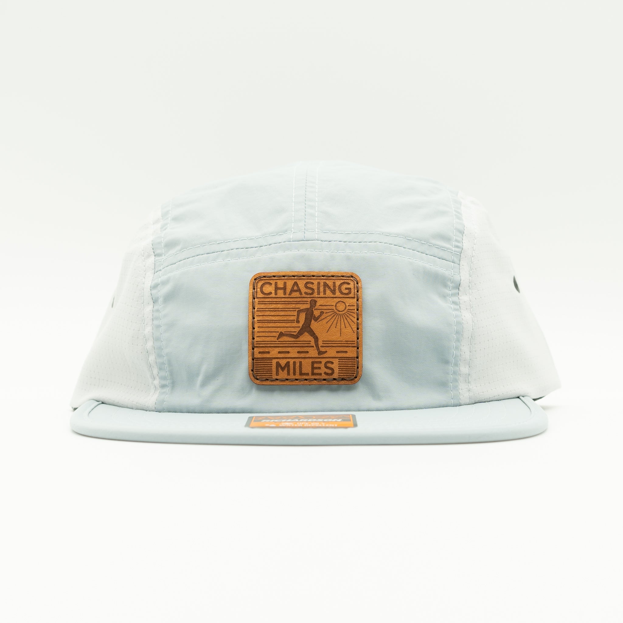 A light blue 5-panel cap with a brown leather patch on the front that reads 'CHASING MILES' with a running figure graphic, photographed on a clean white background.
Color: Grey/Glacier Grey