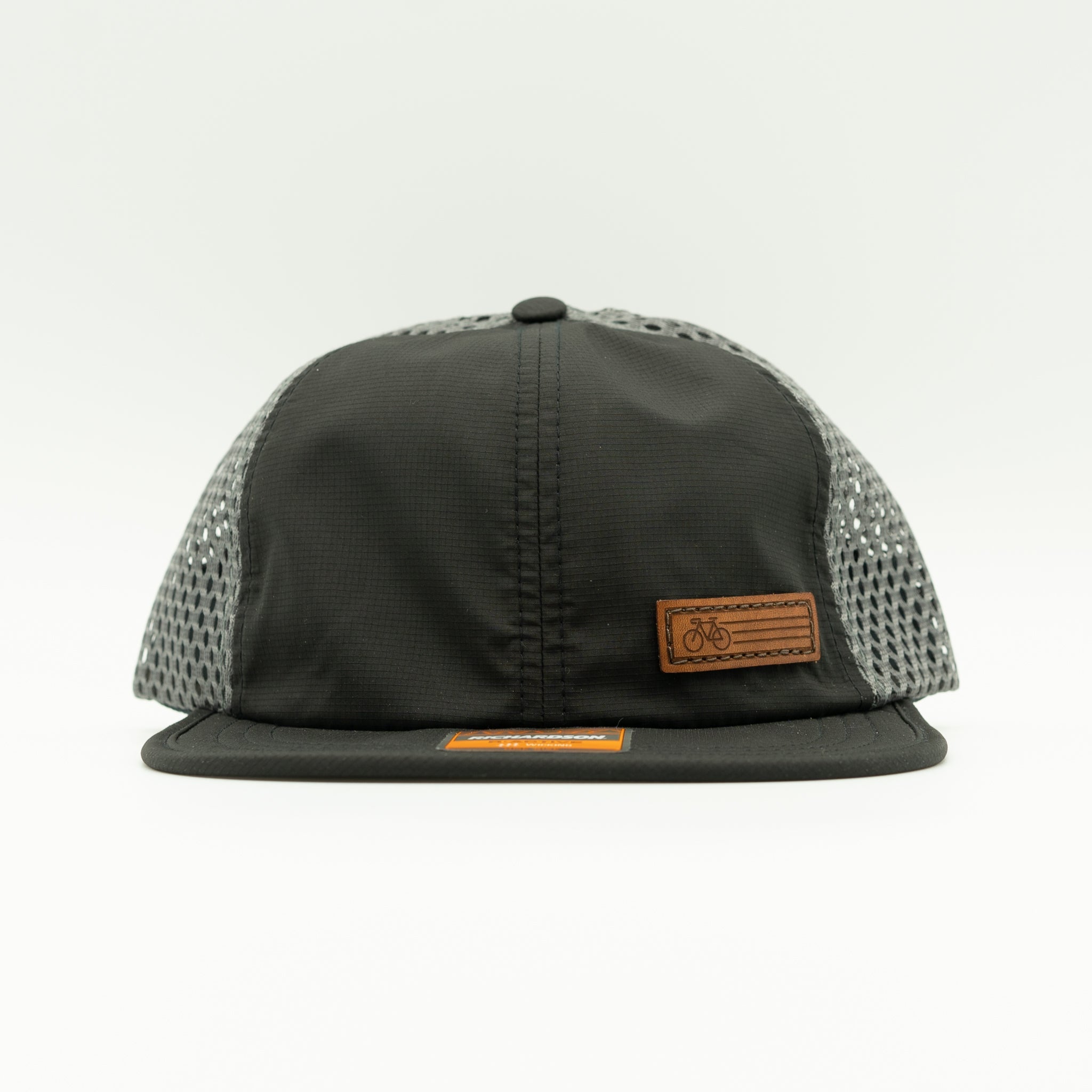 A black trucker-style cap with a black front panel and brim, gray mesh side panels, and two small brown leather patches on the front and brim.
Color: Black/Charcoal