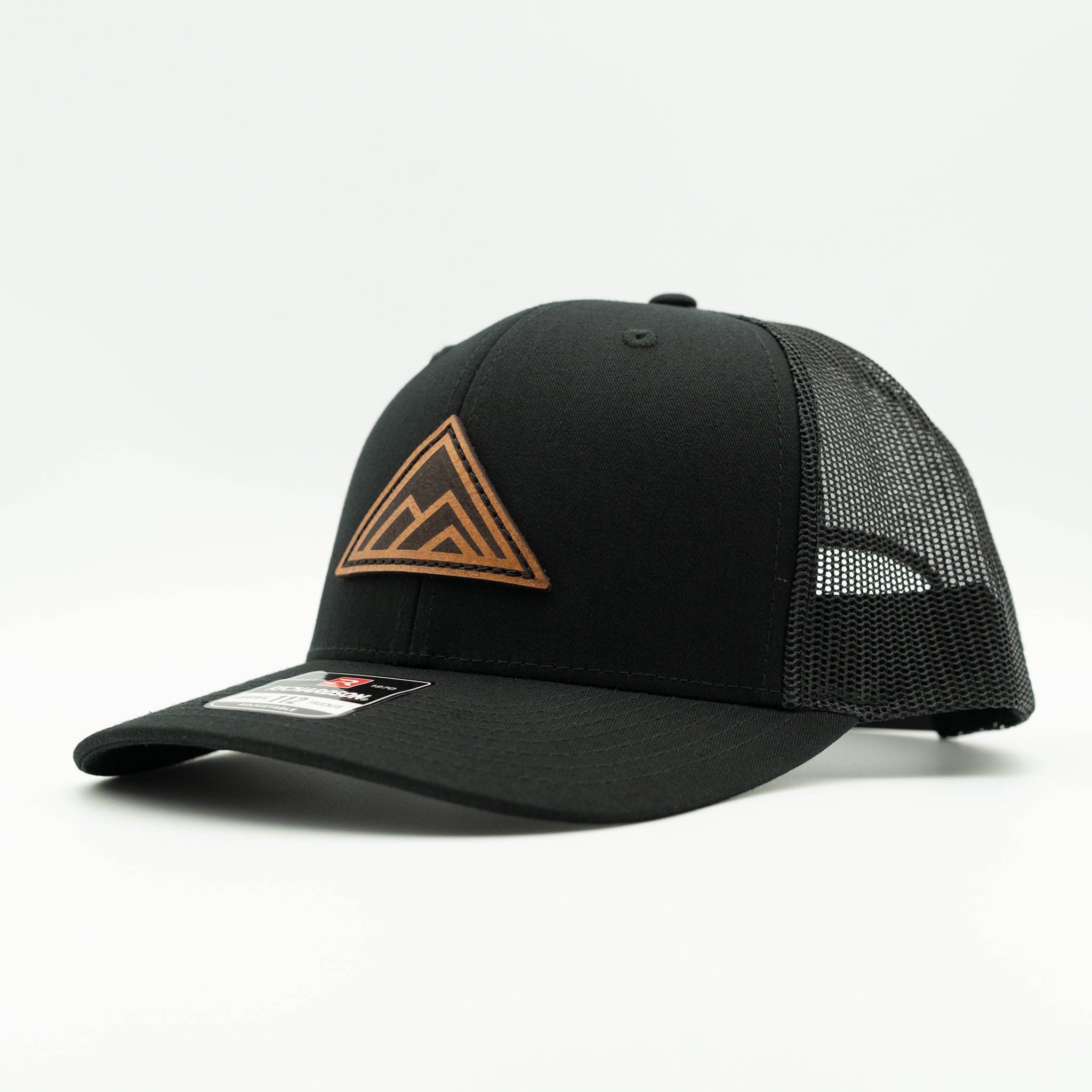 A black trucker-style cap with a curved brim, black mesh back panels, and a brown Leather patch mountain logo on the front, shown against a plain white background.

Color: Solid Black