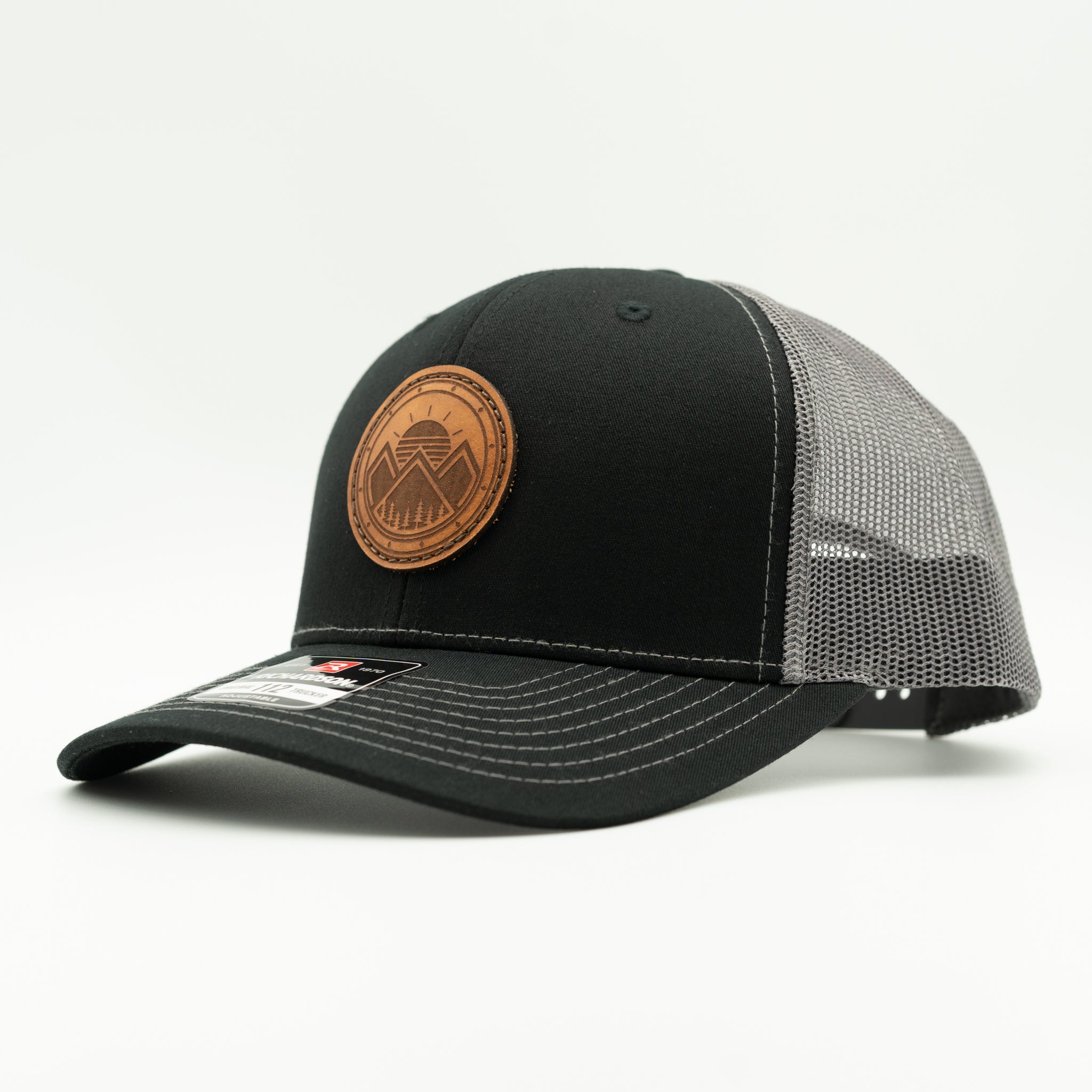 A black trucker-style cap with a curved brim, black front panels, and a gray mesh back. It features a circular brown leather patch on the front with a mountain logo and text, and a small white sticker on the brim.

Color: Black/Charcoal