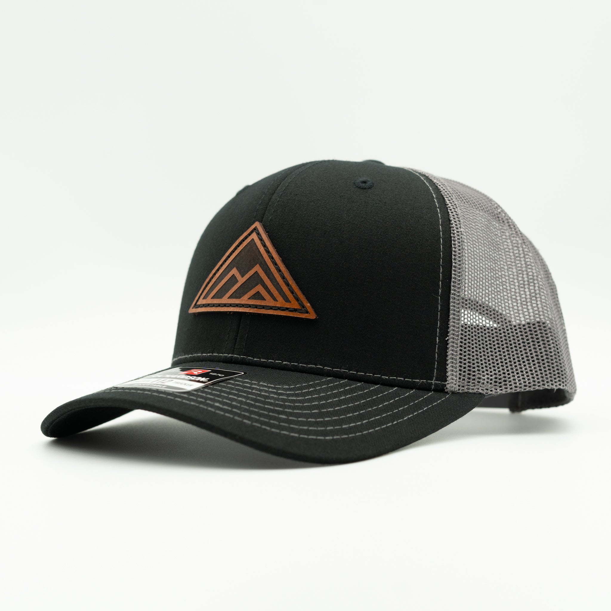 A black trucker-style cap with a curved brim, black front panel, and gray mesh back panels. It features a brown leather patch mountain logo on the front and white contrast stitching on the brim.

Color: Black/Charcoal