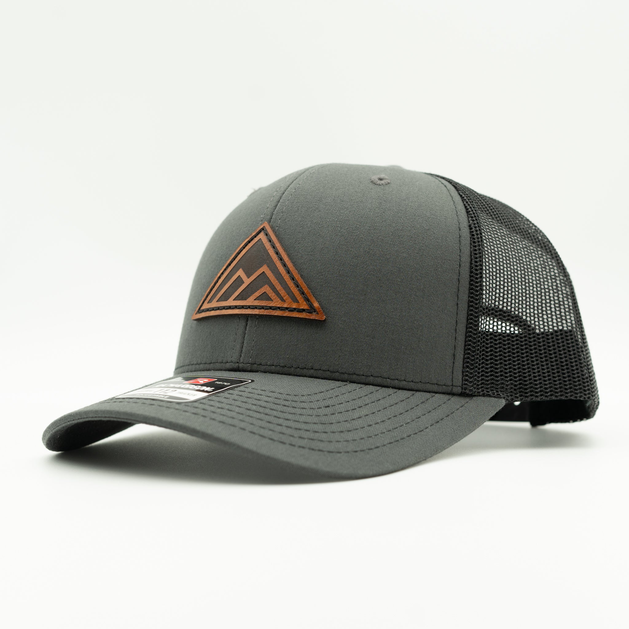 A dark gray trucker hat with black mesh back panels and a brown geometric mountain logo patch on the front, shown on a light gray background.

Color: Charcoal/Black