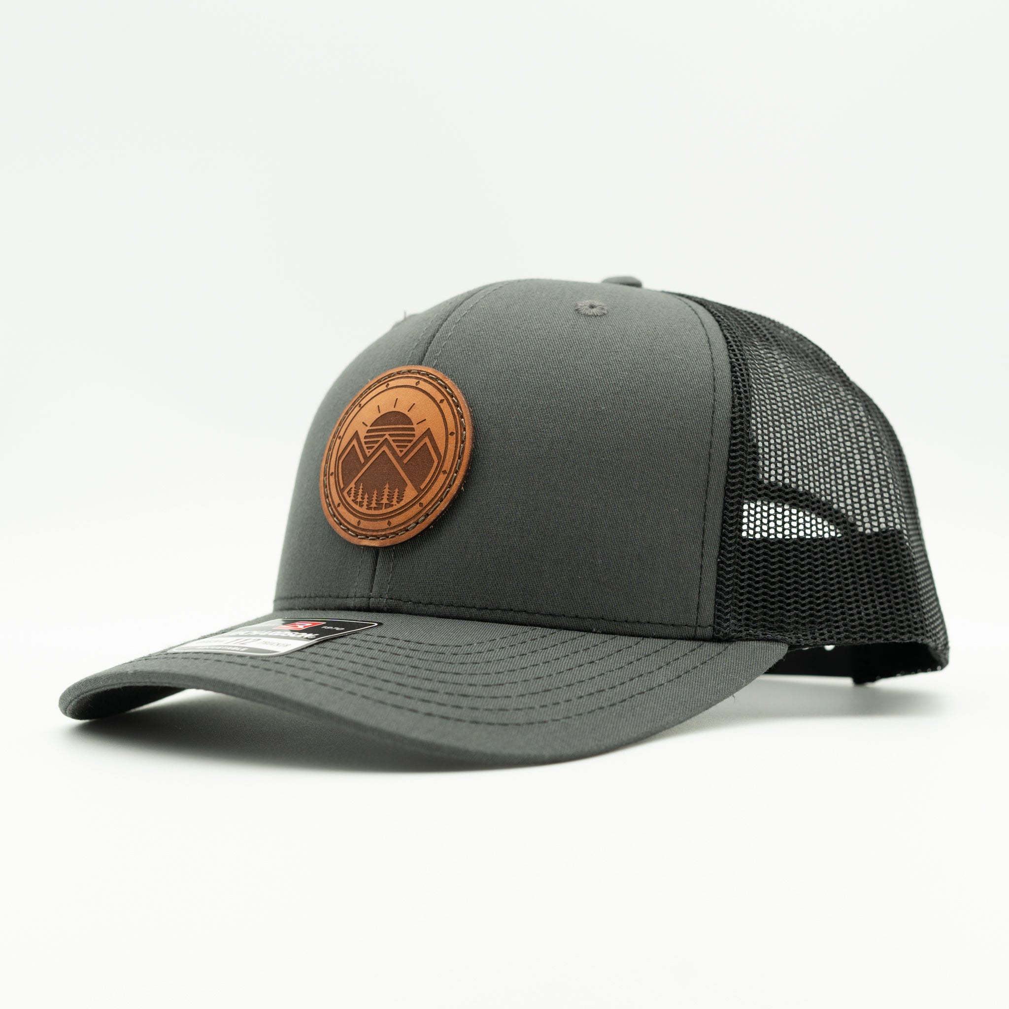 A dark gray trucker hat with a black mesh back and a circular brown leather patch on the front featuring a stylized eagle logo.

Color: Charcoal/Black