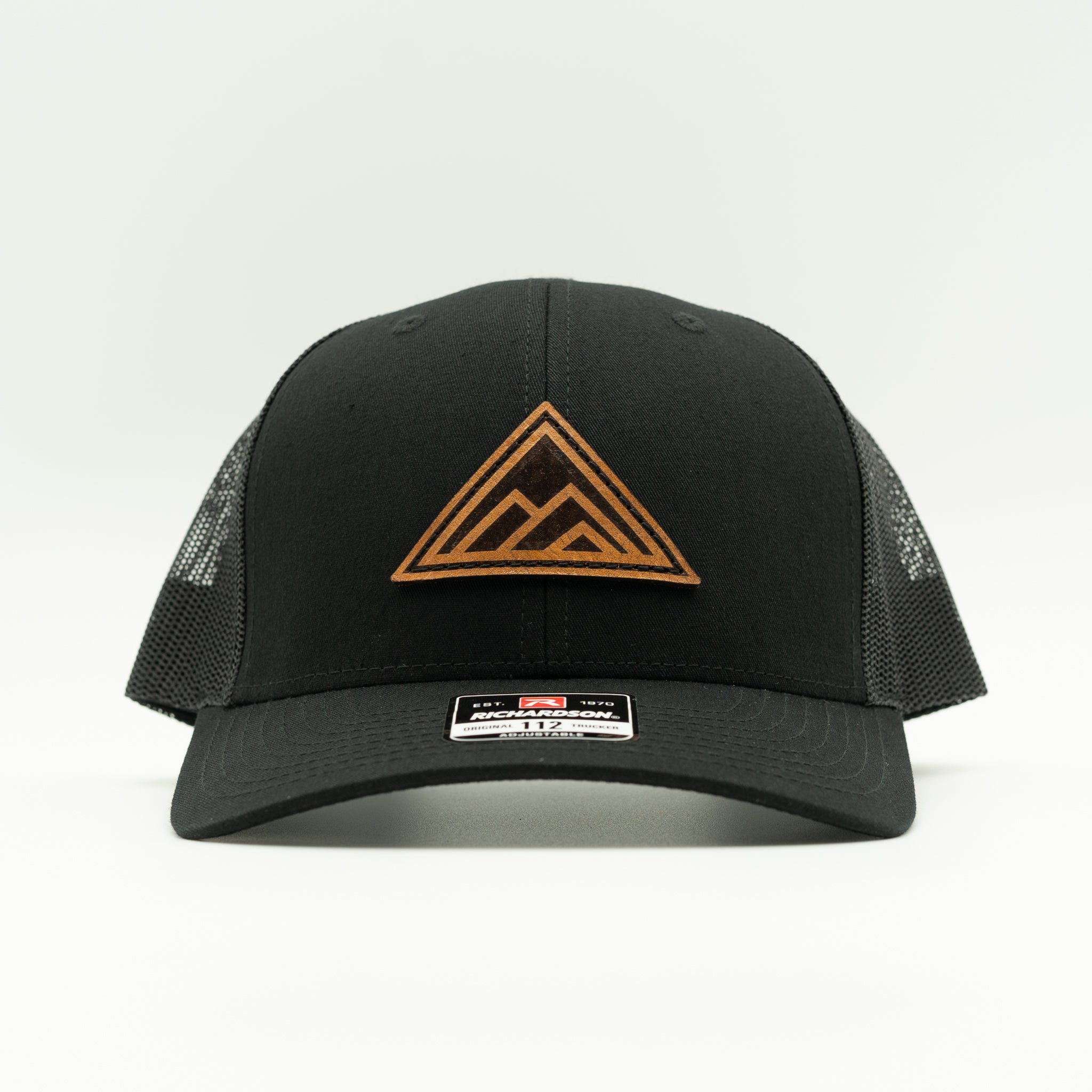 A black trucker-style cap with a brown leather patch mountain logo on the front panel and a small brand patch on the brim, photographed against a clean white background.

Color: Solid Black