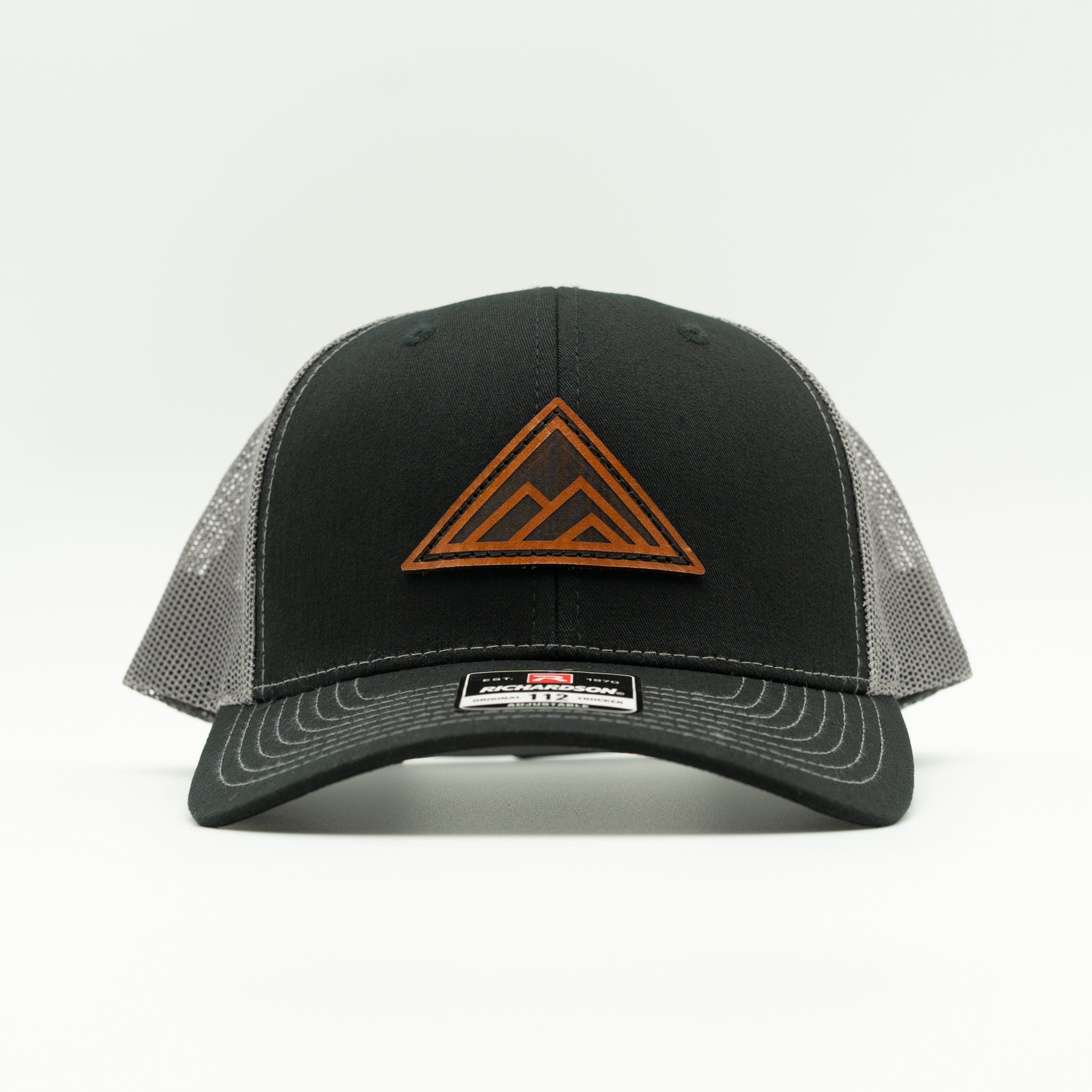 A black trucker-style baseball cap with a brown leather patch mountain logo on the front and gray mesh side panels, shown against a plain white background.

Color: Black/Charcoal