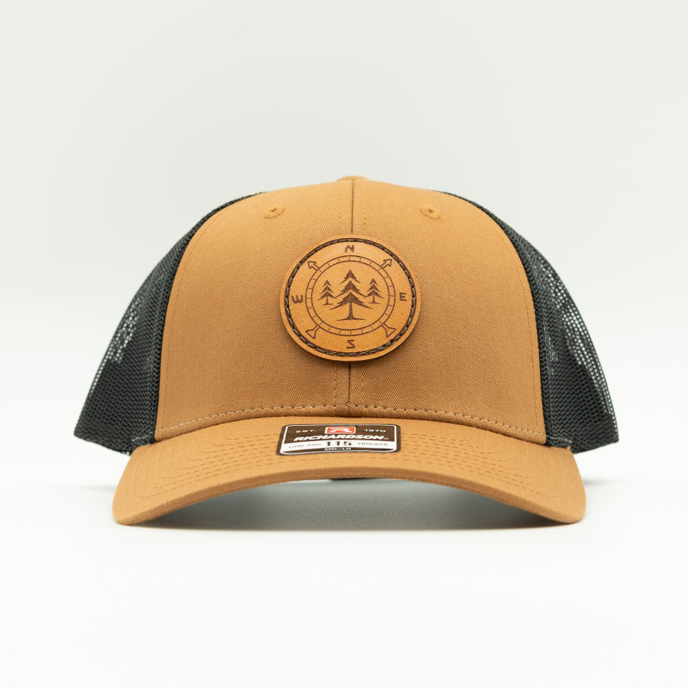 Front view of the Lost Pine Hat in Caramel/Black, showcasing the breathable mesh back, structured profile, and adjustable closure. The hat includes a hand-stitched leather patch with a compass and pine trees design, perfect for those who love the outdoors.