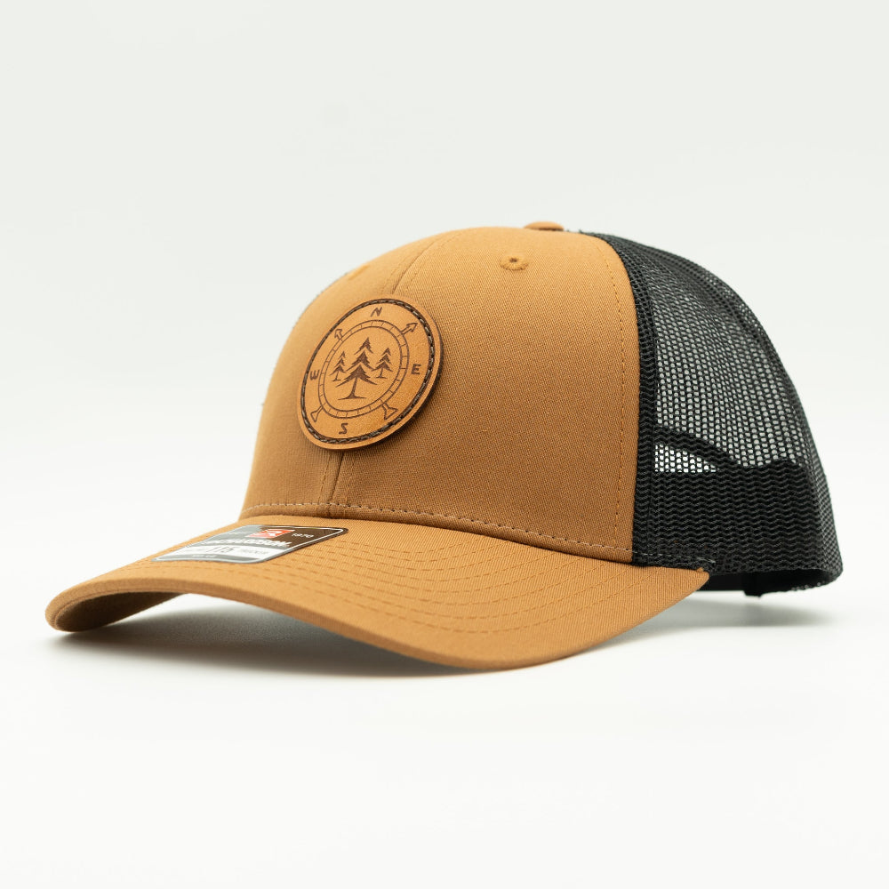 Side view of the Lost Pine Hat in Caramel/Black, showcasing the breathable mesh back, structured profile, and adjustable closure. The hat includes a hand-stitched leather patch with a compass and pine trees design, perfect for those who love the outdoors.