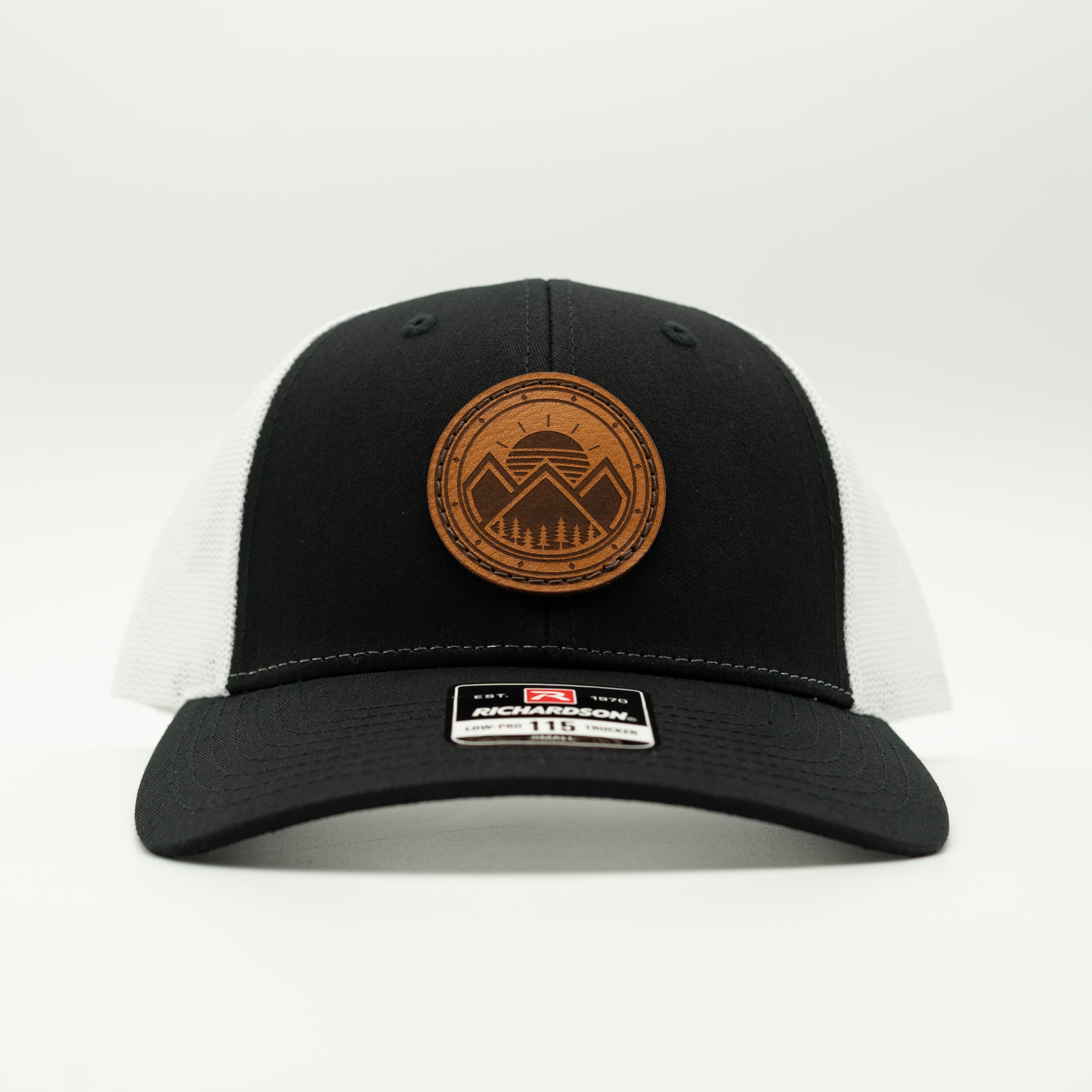 A black and white trucker hat with a brown circular leather patch on the front. The patch features a mountain and sun design with the text. 

Color: Black/White