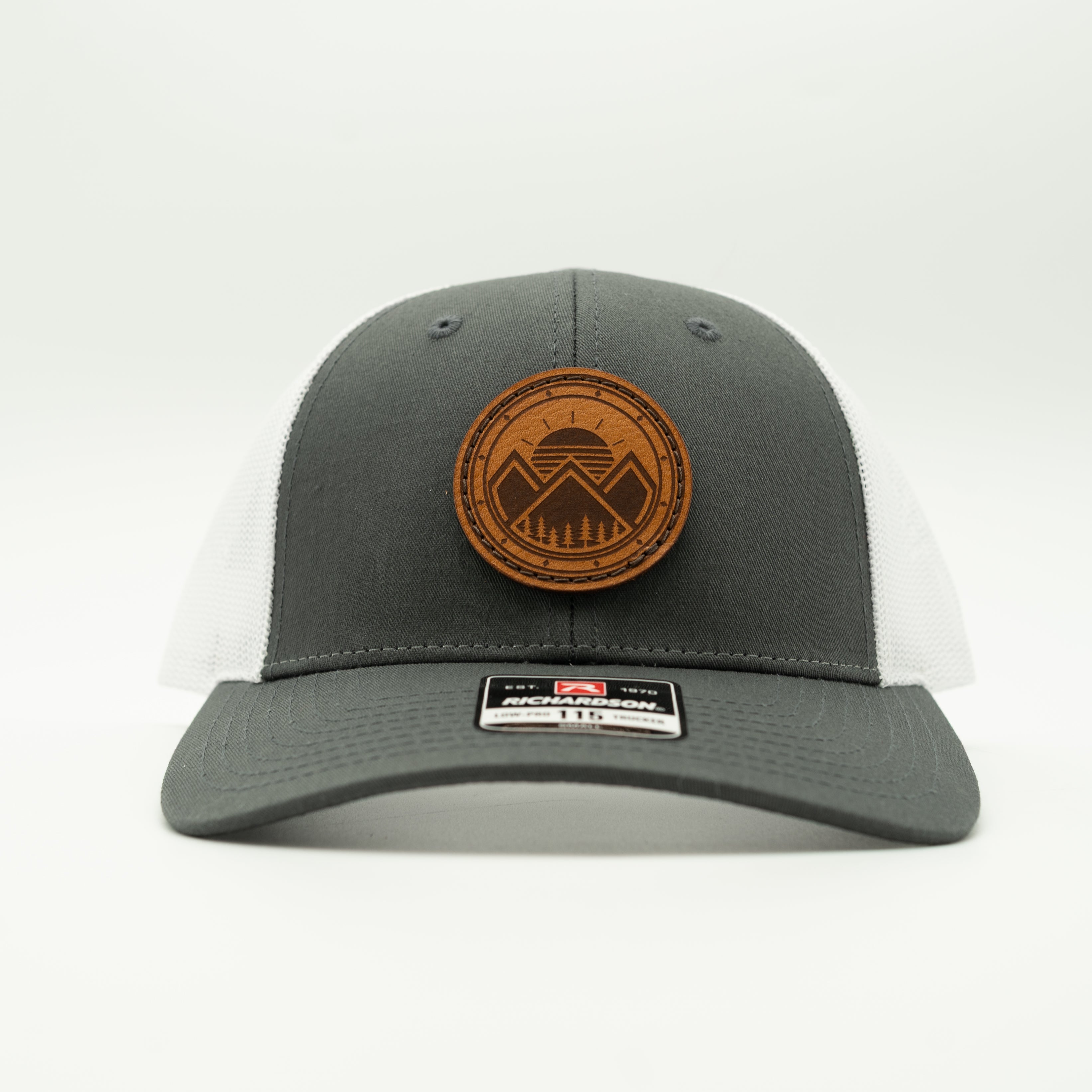 A dark gray and white trucker hat with a brown leather patch on the front displaying a mountain logo. The hat features a dark gray front panel and brim, white mesh back panels.

Color: Charcoal/White