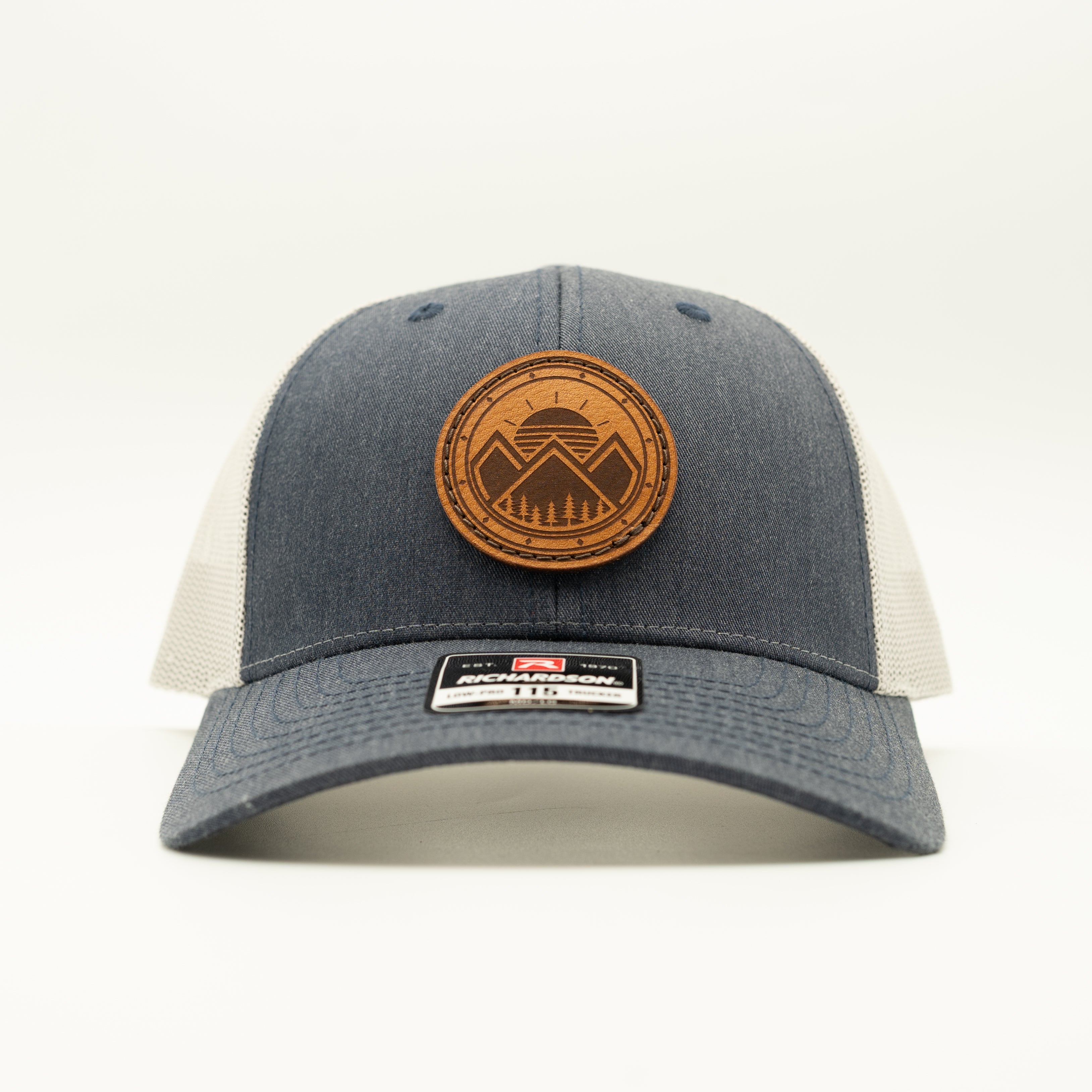 A dark gray and off-white trucker hat with a circular brown leather patch on the front displaying a mountain logo.

Color: Heather Navy/Grey
