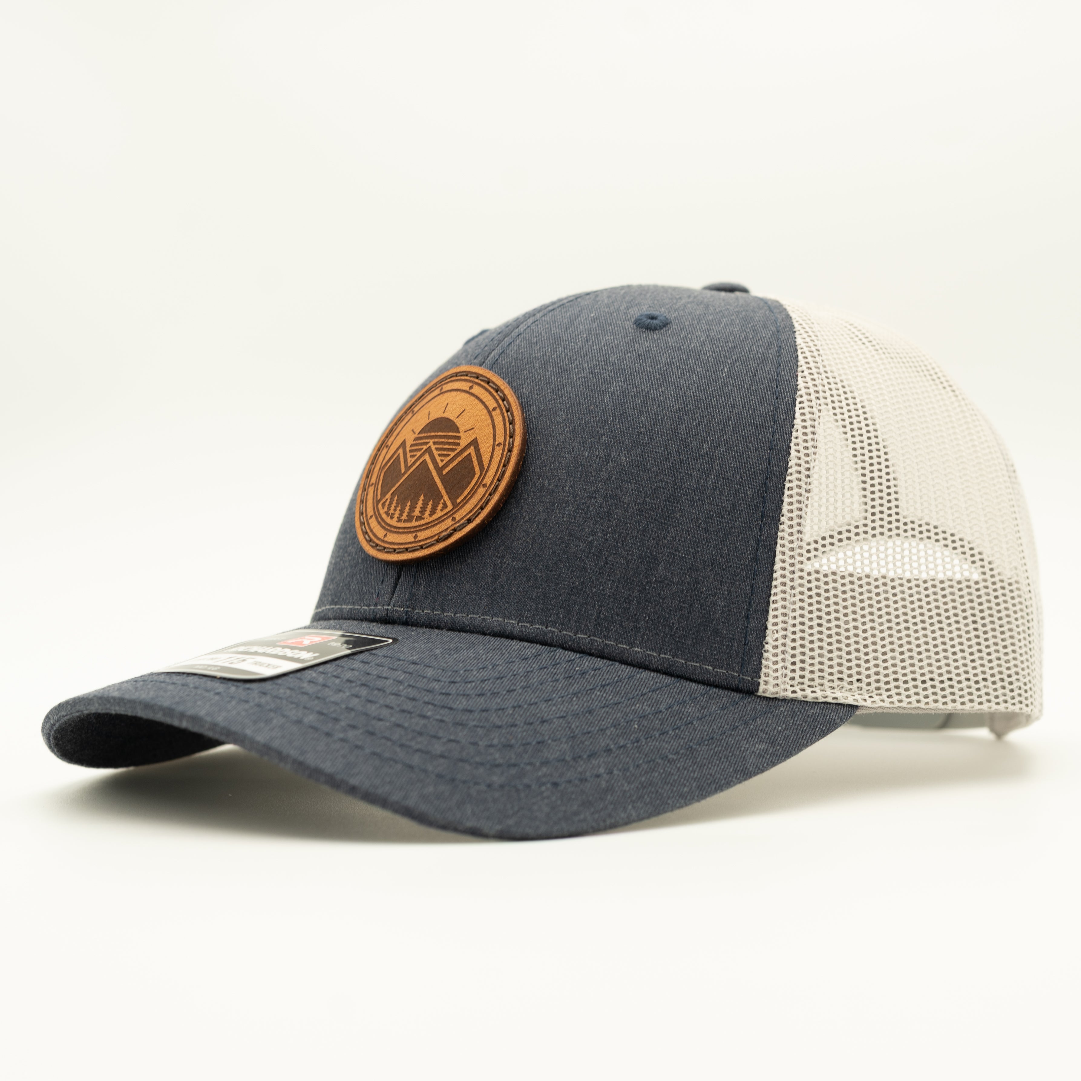 A trucker-style baseball cap with a dark heather gray front and visor, cream mesh back panels, and a circular brown leather patch on the front.

Color: Heather Navy/Grey