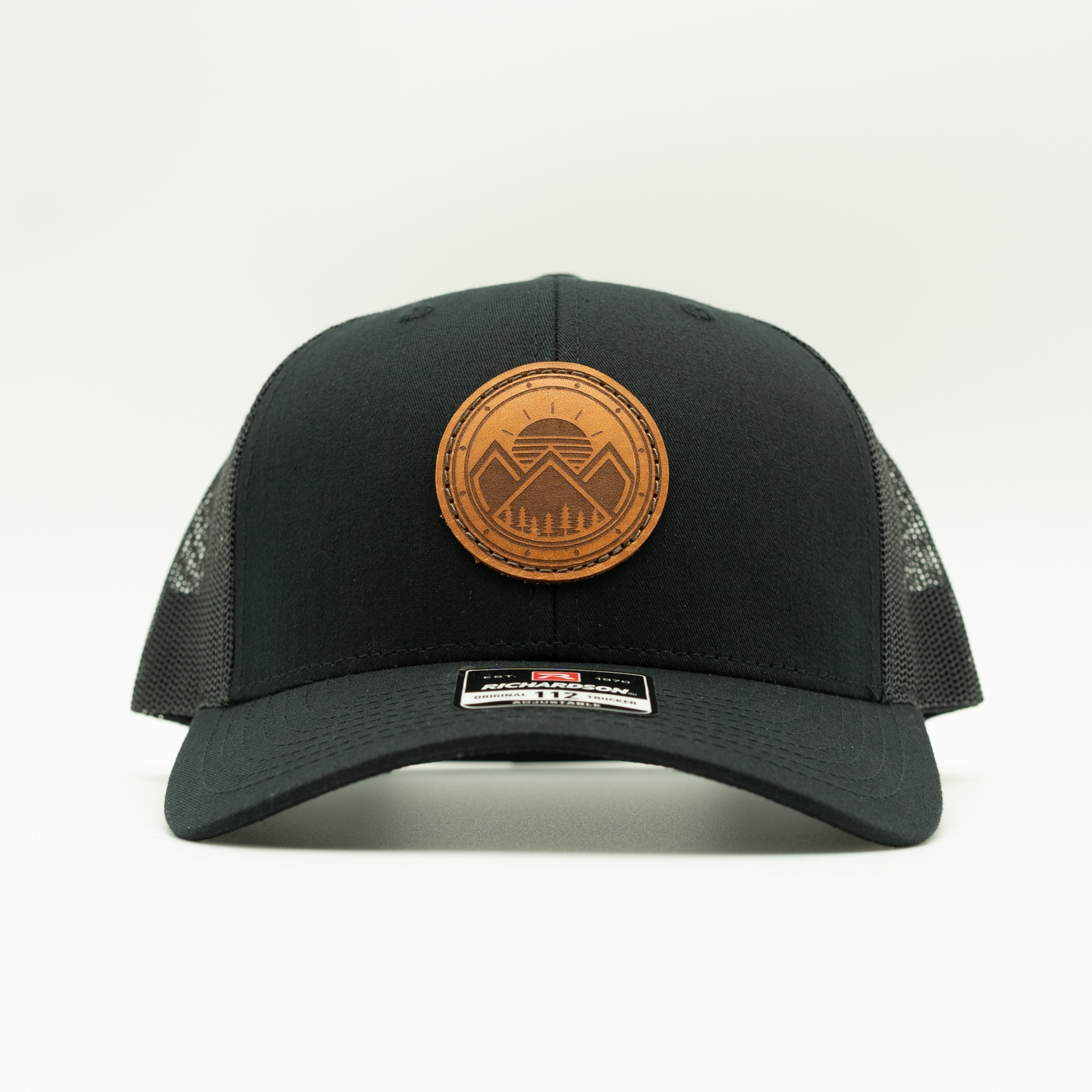 A black trucker hat with a brown circular leather patch on the front featuring a mountain and dome design. The hat has black mesh side panels and a black curved brim, photographed against a clean white background.

Color: Solid Black