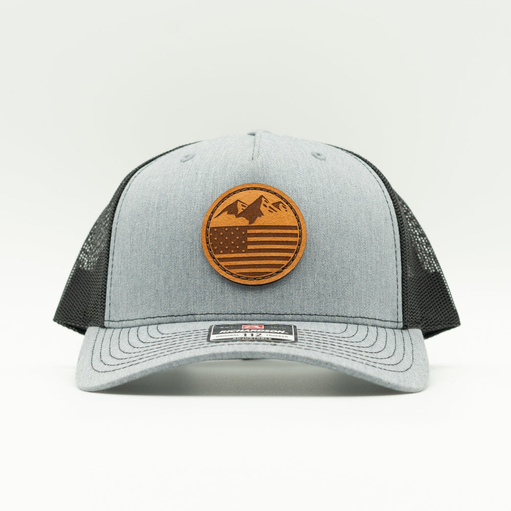 Front view of the Mountain Flag leather patch hat on Richardson 112FP in Heather/Black, highlighting the flat bill, adjustable snapback, and seamless front panel. The perfect custom leather patch hat for outdoor activities.