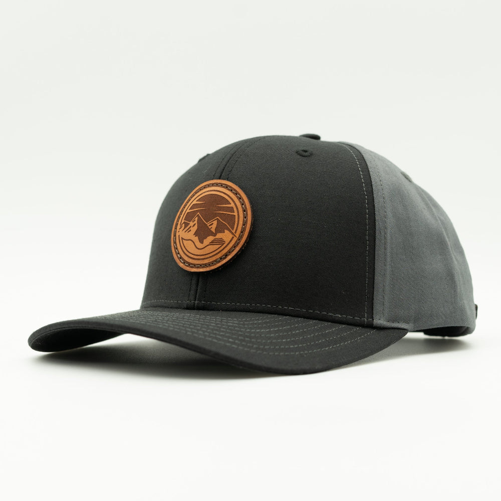 Side view of the Mountain Lake leather patch hat on Richardson 312 in Black/Charcoal, highlighting the pre-curved visor, adjustable snapback closure, and full-fabric front. A perfect custom leather patch hat for mountain man apparel.